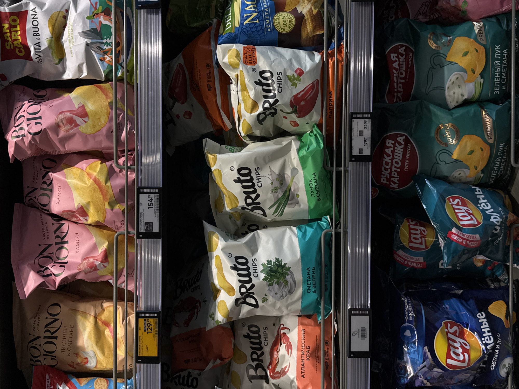 The image shows a shelf in a Moscow, Russia supermarket filled with various bags of potato chips. The chips are arranged on three tiers of shelving. The top shelf displays bags from "Bon Giorno" and "San Carlo" brands, featuring flavors like "Lichichi v Smetane" (Crayfish in Sour Cream), "Khamon" (Jamon), and "Lime & Pink Pepper". The middle shelf is dominated by "Bruto Chips" with flavors such as "Atlantichveskiy Krab" (Atlantic Crab), "Smetana & Zelen" (Sour Cream & Herbs), and "Paprika". The bottom shelf features "Lay's" chips in packaging for "Fantastika Bkcheec Lay" and "Smetana i Luk" (Sour Cream and Onion), as well as "Russkaya Kartoshka" (Russian Potato) chips in "Zelyonyi Luk i Smetana" (Green Onion and Sour Cream) flavor. Prices are visible on electronic shelf labels for some of the products. The lighting suggests an indoor commercial environment. There are no people visible in the image.