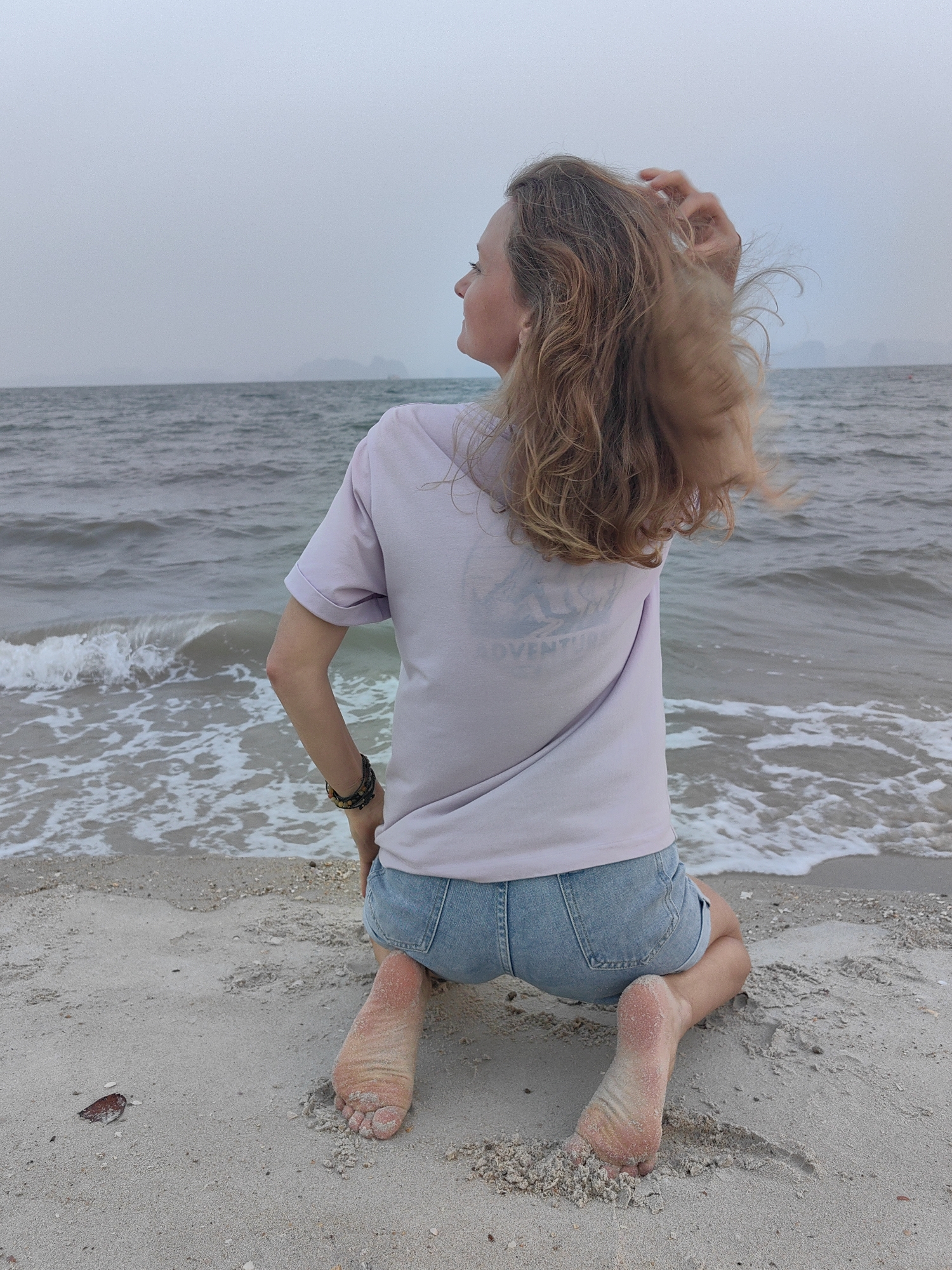 A woman is kneeling on a sandy beach at Bai Chay Ward, Vietnam, facing away from the viewer and toward the water. She has fair skin and long, wavy, light brown hair. She is wearing a light purple T-shirt with a faded graphic on the back and denim shorts. Her right hand is raised and positioned in her hair. Her bare feet are in the sand. The water is grayish-blue and contains white foamy waves. In the distance, there are some land formations on the horizon, silhouetted against an overcast sky.