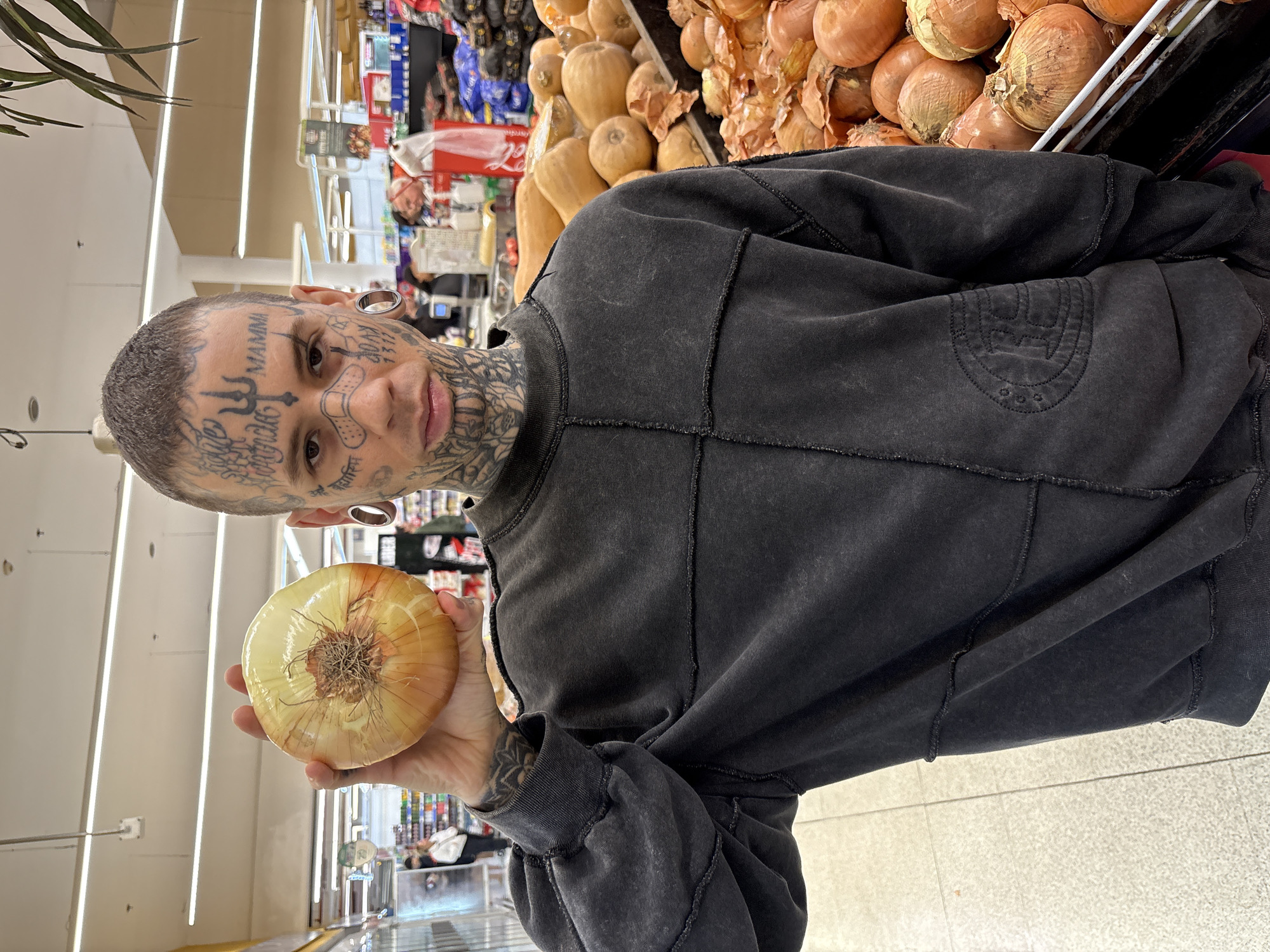 The image depicts a person in a supermarket produce section, likely in Buenos Aires, Argentina, holding up a large onion towards the camera. The individual is characterized by extensive tattoos covering their face, neck, and arms, and wears large ear gauges. Their hair is short and dark.
Prominent facial tattoos include the word "MAMMA" on their forehead, a band-aid design tattooed across their nose, and intricate patterns around their eyes, cheeks, and jawline, extending down their neck. They are wearing a dark grey or black long-sleeved sweatshirt that appears worn or distressed, with visible stitched seams and a subtle embossed logo on the chest.
The person's right hand holds a single, light-skinned onion, presented prominently. In the background, large bins are filled with numerous similar onions, along with other produce like butternut squash. Further back, supermarket aisles with various packaged goods and overhead fluorescent lighting are visible. Faintly, a "Coca-Cola" branding can be seen on a red display in the distance. The lighting is bright and even, typical of an indoor retail environment. The person looks directly at the camera with a neutral expression.