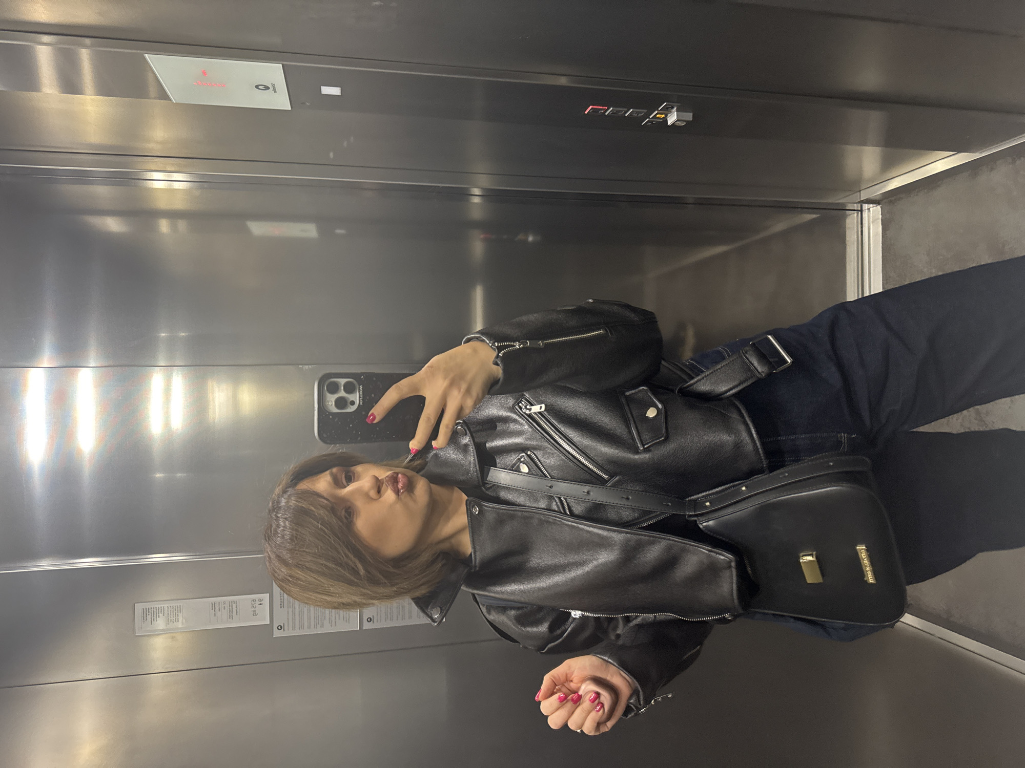 The image captures a young woman taking a selfie in an elevator. She is wearing a black leather jacket over dark clothing and has a black shoulder bag. Her hair is shoulder-length and brown, and she has red nail polish. The elevator is metallic and modern, with signs and buttons visible on the walls. The location is uncertain, and there is no explicit information to connect this image to Sofia, Bulgaria.