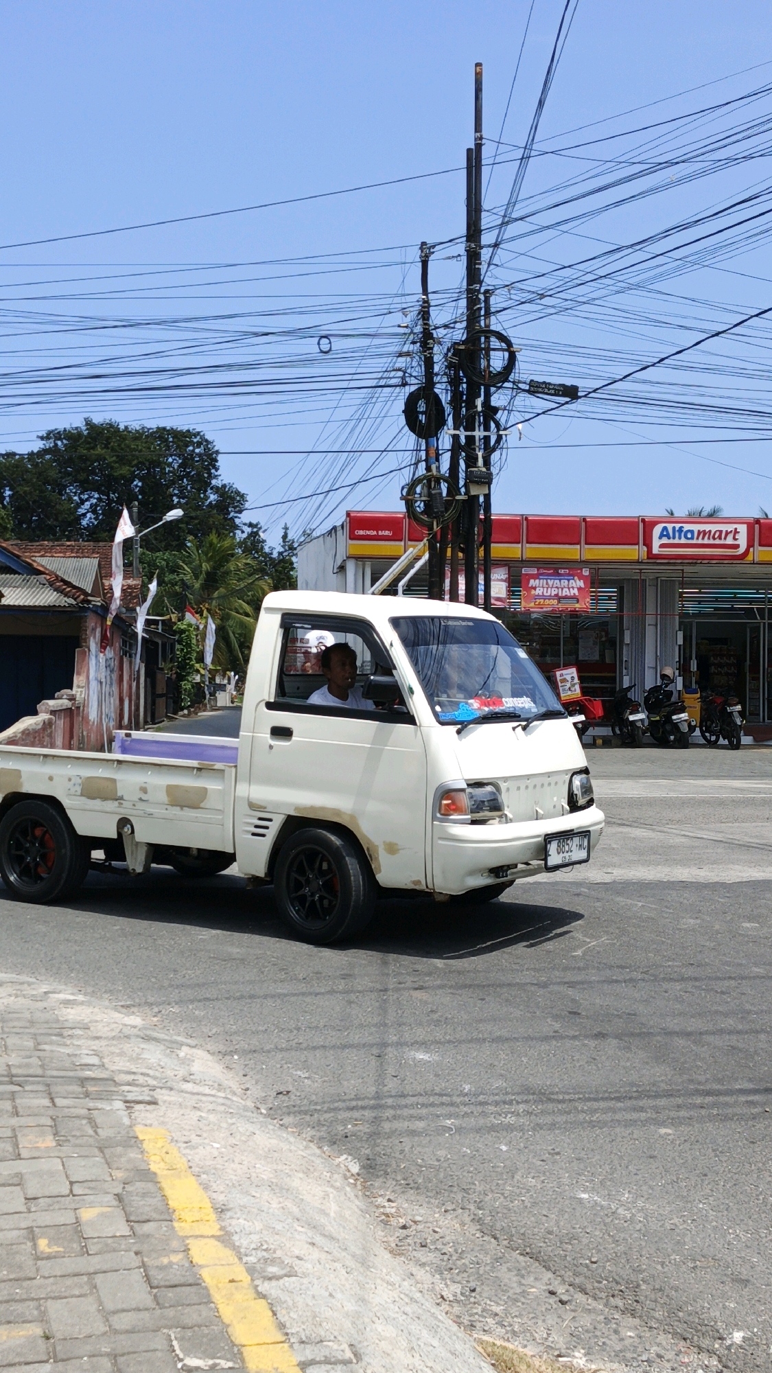 A white pickup truck drives down a paved road on a sunny day in Cibenda, Indonesia. The truck has a driver in the cab. In the background, there is an Alfamart convenience store, a utility pole with many wires, and buildings. The sky is clear and blue.