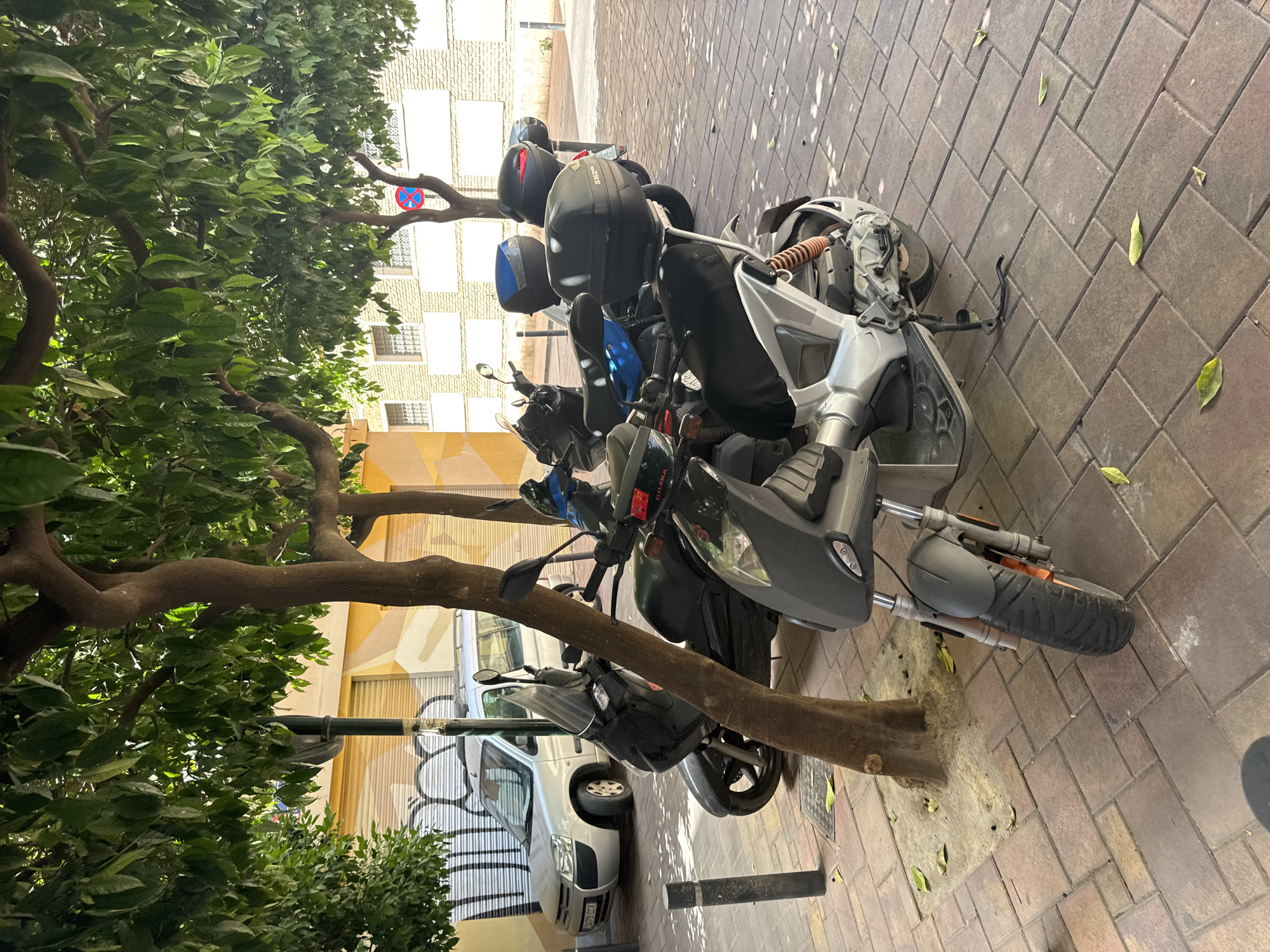 The image depicts a street scene in Murcia, Spain, characterized by a row of parked scooters and a silver car. The foreground is dominated by a silver and gray scooter with an orange suspension component, parked on a patterned brick sidewalk. Behind it, several other scooters are visible, some with storage boxes on the back, and a silver car is parked further down the street. The scooters and car are parked beneath trees with dense green foliage that casts dappled shadows on the ground. In the background, buildings with closed shutters and window details are visible. A street sign with a red circle and a blue "X" is also present. The lighting suggests it is daytime.