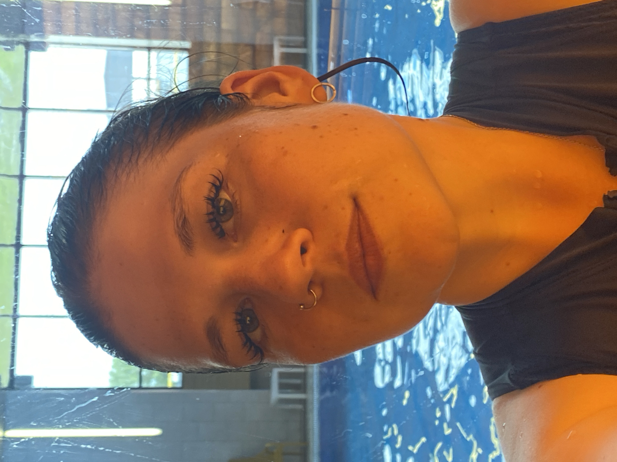 A woman with dark hair, wet from swimming, is captured in a close-up selfie. Her eyes are open and looking forward, and she has a nose ring and hoop earrings. The background shows a swimming pool with clear blue water, and large windows that reveal a glimpse of the outside world, possibly Votkinsk, Russia, given the reference. The lighting is warm, casting an orange hue over the scene. She is wearing a dark-colored top.
