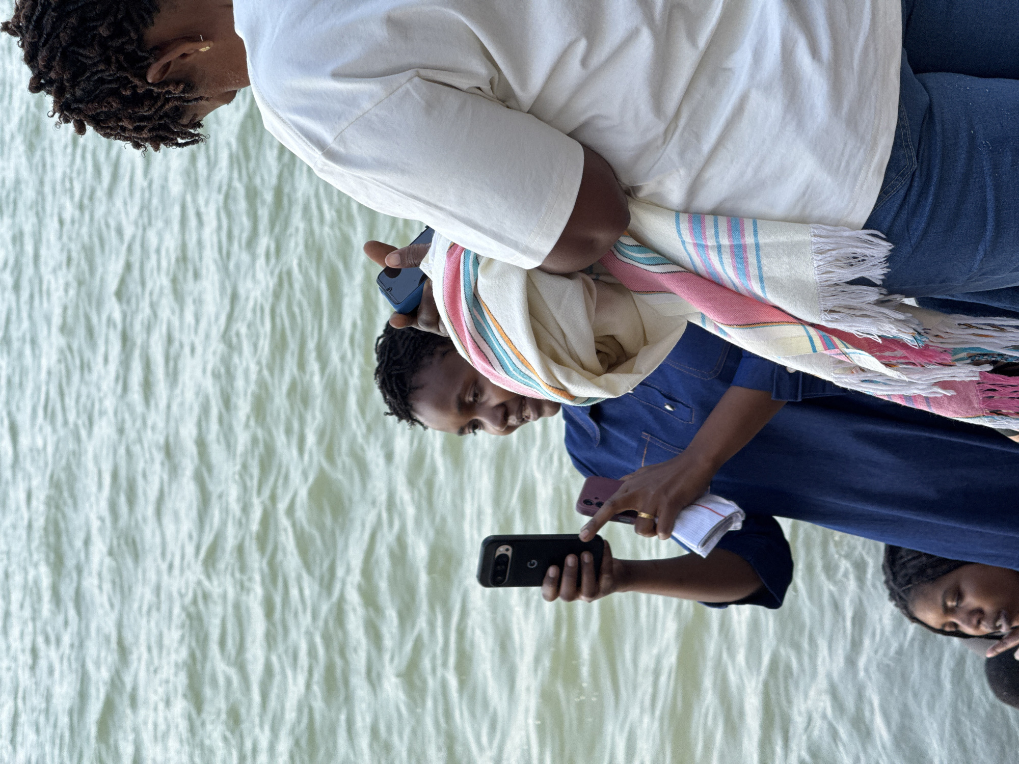 In Dar es Salaam, Tanzania, two individuals are captured near the ocean. The person on the left, wearing a denim shirt, holds two phones, one black and one purple, in their hands, appearing to be taking a photo or recording a video. They are standing in front of a person in the water. The person on the right, wearing a white t-shirt and jeans, is holding a brightly colored striped towel. The water is choppy, suggesting a breezy day. The scene is candid and appears to be a moment of casual photography by the sea.