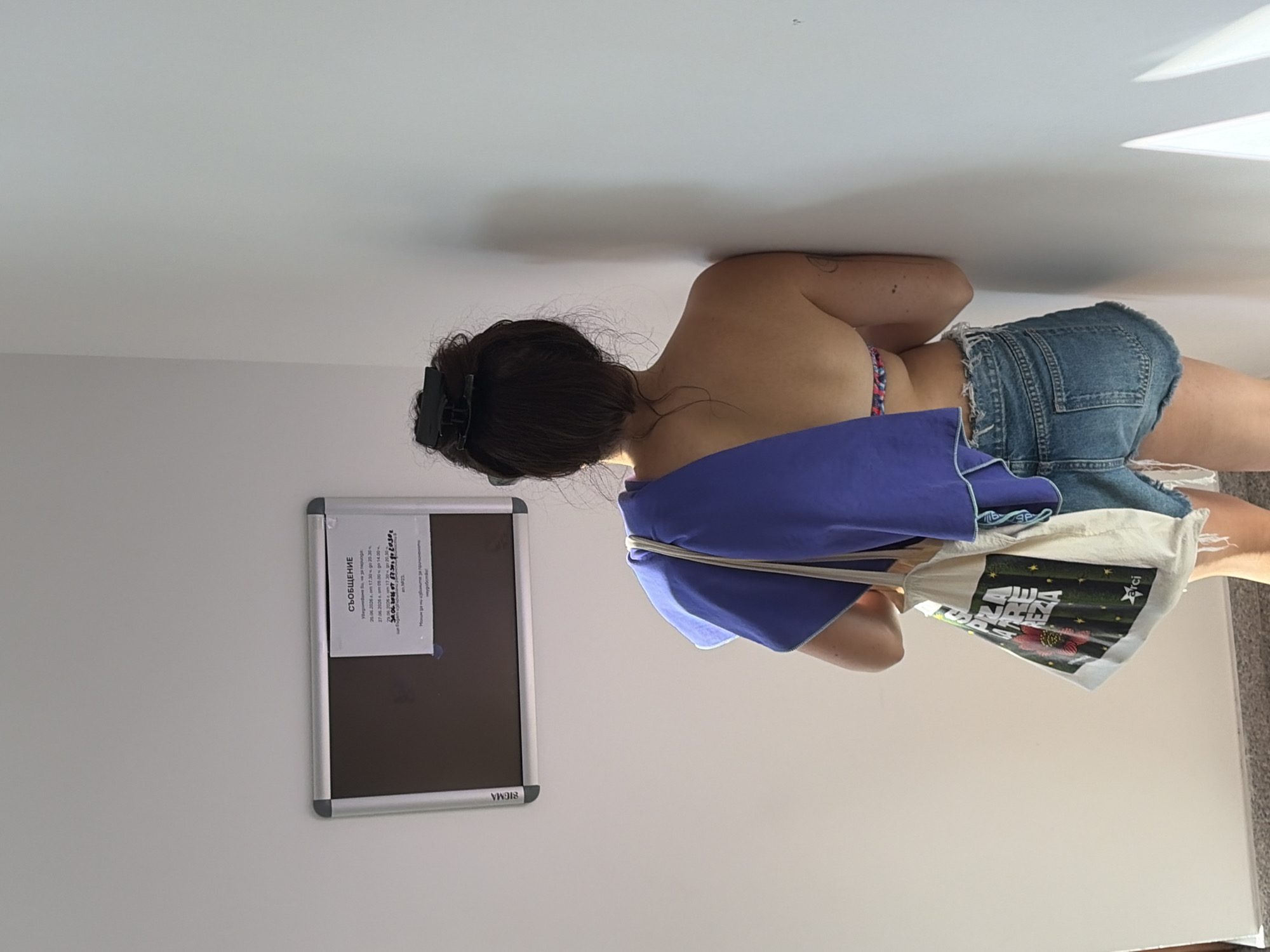 A person is captured in a vertical, low-angle shot, leaning against a white wall. They are wearing denim shorts and a colorful bikini top, with a purple cloth draped over their shoulder and a canvas tote bag slung over their arm. The tote bag has an image of a red flower and some text. A bulletin board with some text is visible on the wall to the left. The lighting suggests it might be daytime, with soft shadows on the wall. The person's hair is dark and tied up, with a clip holding it in place. The image is taken indoors, possibly in a home or studio setting. The visible text on the bulletin board includes "СЪОБЩЕНИЕ" (Announcement), dates and times, and some numbers and letters. The tote bag also has some visible text, including "rci" and what appears to be a stylized logo.