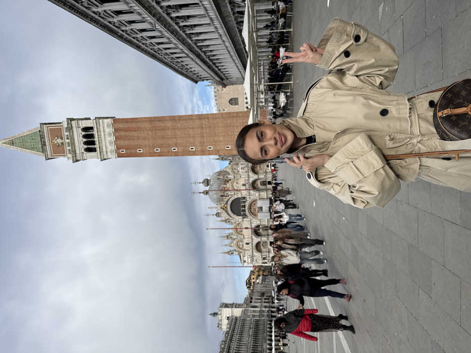 The scene depicts St Mark's Square (Piazza San Marco) in Venice, Italy, under an overcast sky. In the lower right foreground, a woman with dark hair wears a light beige cropped trench coat and a round, brown cross-body bag with a monogram pattern. She holds a silver smartphone to her right ear with her right hand, appearing to be on a call, while simultaneously displaying a "peace" sign with her raised left hand and smiling towards the viewer.
Behind her, in the central background, stands St Mark's Basilica, characterized by multiple domes, arches, and an ornate facade. To the right of the Basilica, St Mark's Campanile, a tall brick bell tower with a green pyramidal spire, dominates the skyline. Adjacent to the Campanile, a grand classical building with multiple levels of arched windows borders the square, in front of which an outdoor cafe with white umbrellas, tables, and seated patrons is visible. In the mid-ground, a line of approximately 20-30 individuals walks from right to left across the square. The ground of the piazza is paved with light-colored square tiles.