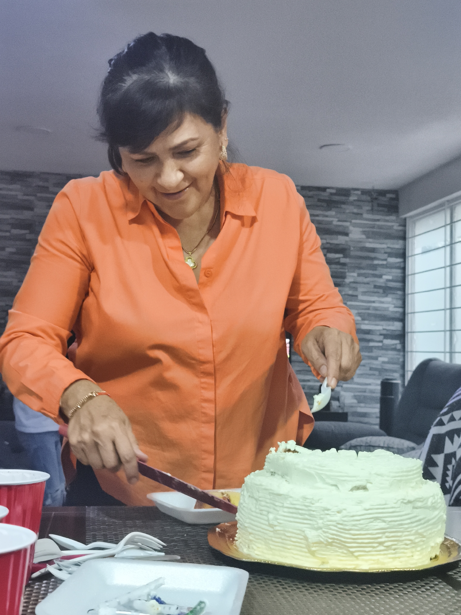 A woman is cutting a two-tier cake on a table. The cake has white frosting. The woman is wearing an orange button-down shirt, a necklace, and a bracelet. Red cups and white plastic utensils are on the table near the cake. A window is visible in the background. The location is unclear, and I cannot verify that the location is Mexico City.