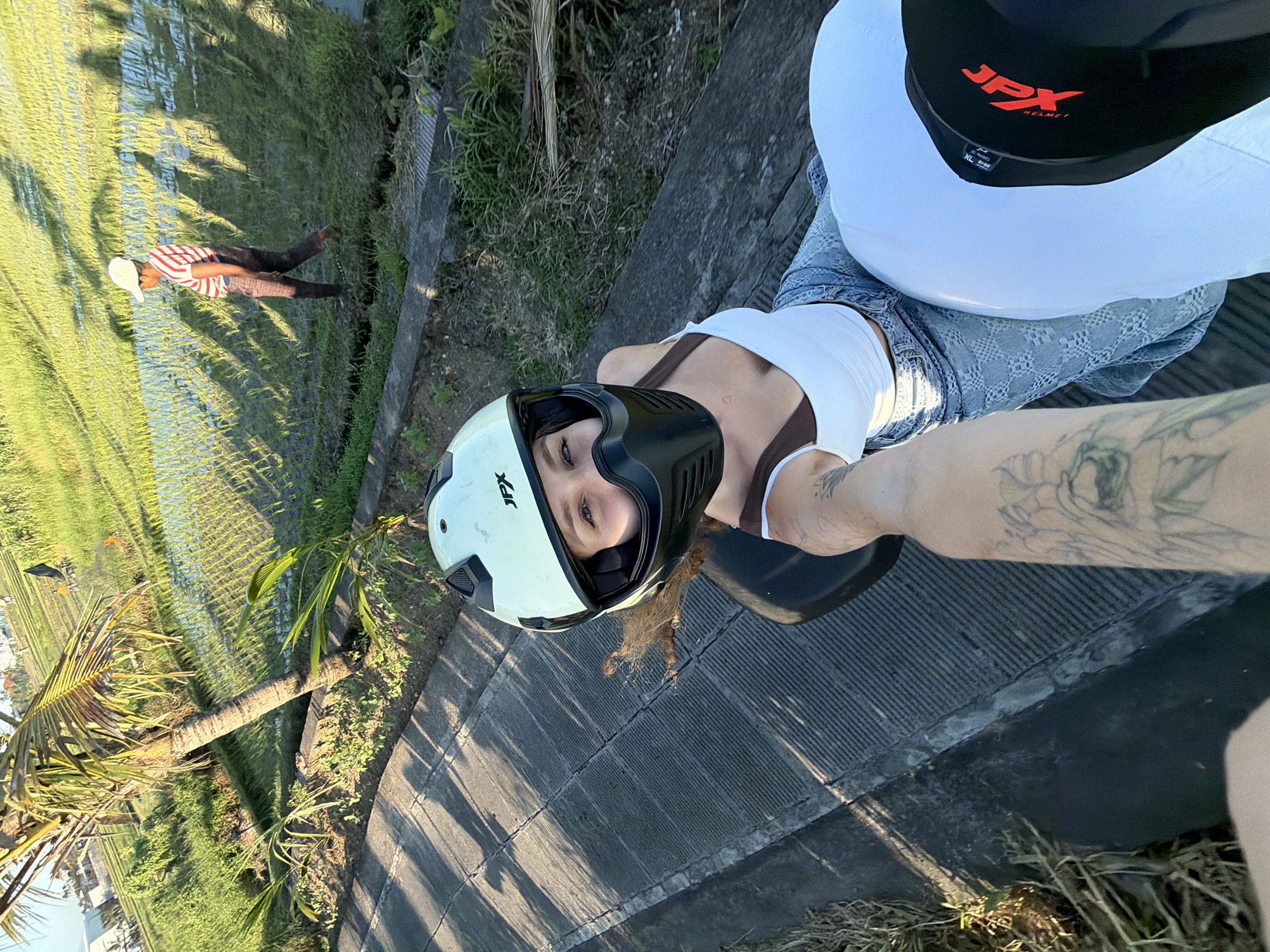 A medium-shot, selfie-style photograph shows a woman wearing a motorcycle helmet, sitting on a scooter on the edge of a rice field in North Kuta, Indonesia. The woman is wearing a white tank top and blue jeans. A black helmet rests on the scooter's seat beside her. In the background, a farmer works in the rice field. A palm tree leans over the edge of the field. The photograph is taken from a high angle, looking down at the woman. The lighting suggests it is either early morning or late afternoon. The overall mood is casual and relaxed.