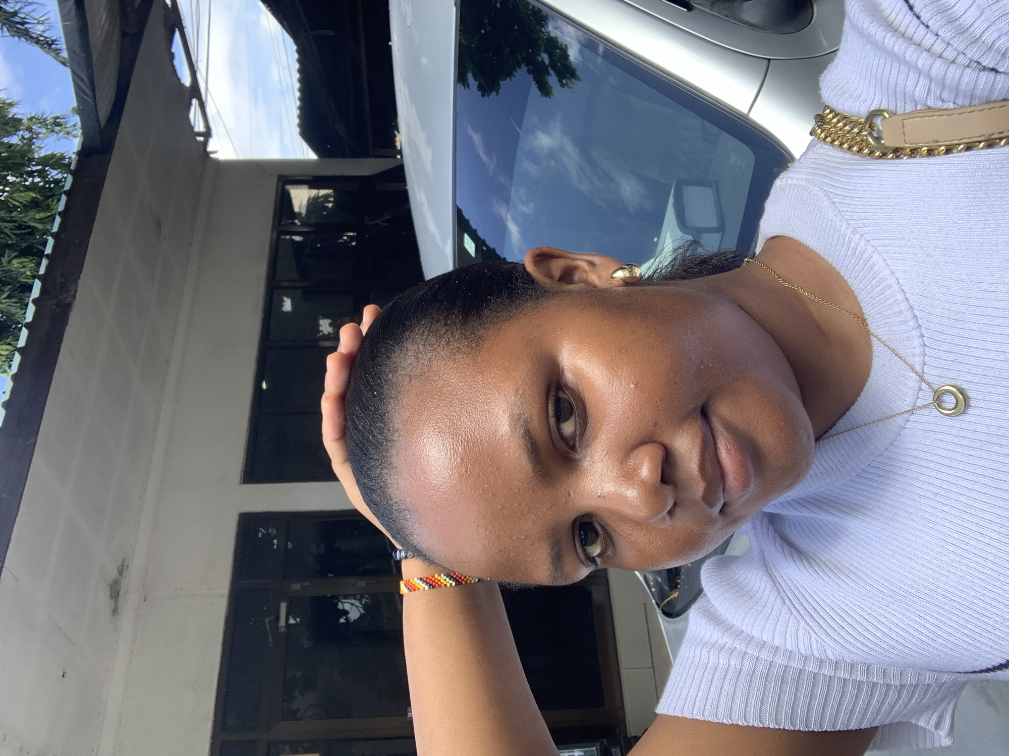 A woman is posing for a selfie, standing next to a silver car. The woman has her hand on her head and is wearing a white shirt, a gold necklace with a ring pendant, and a beaded bracelet. The background includes a building with windows, trees, and a cloudy sky. The reflection in the car window shows the sky and some trees. The location is likely a residential or commercial area in Dar es-Salaam, Tanzania, but the specific location cannot be determined from the image.