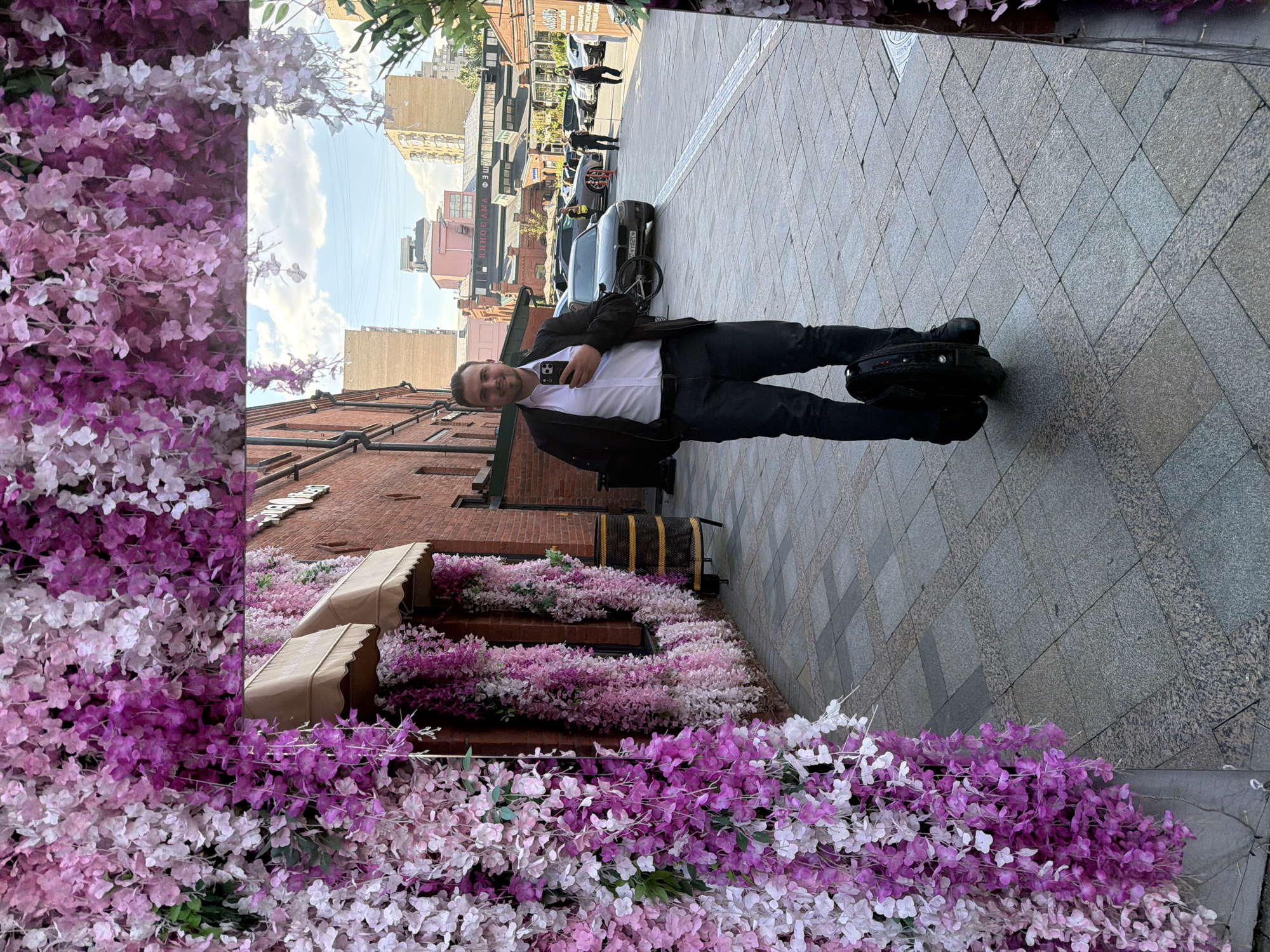 The image displays an urban street scene in Moscow, Russia, during daytime under a clear sky with scattered clouds. A man is the central subject, standing on a grey tiled sidewalk, facing the camera with a slight smile. He has short dark hair and is dressed in a white collared shirt, a dark jacket, dark trousers, and dark shoes. He holds a smartphone in his left hand. Next to his right foot rests a black self-balancing electric unicycle.
To the left of the man, and extending along the foreground, is an extensive and dense display of artificial flowers, primarily in shades of purple, lavender, and white, forming a decorative barrier or wall. Similar floral arrangements adorn the ledges of a multi-story brick building directly behind the man, which also features several brown awnings.
In the background, further down the paved street, more urban buildings are visible, alongside several parked cars and a bicycle. The bright natural lighting suggests good weather. Some Cyrillic text is faintly visible on distant buildings and signs, but it is too far or too blurry to be clearly legible. The overall impression is a pleasant, decorated street environment.