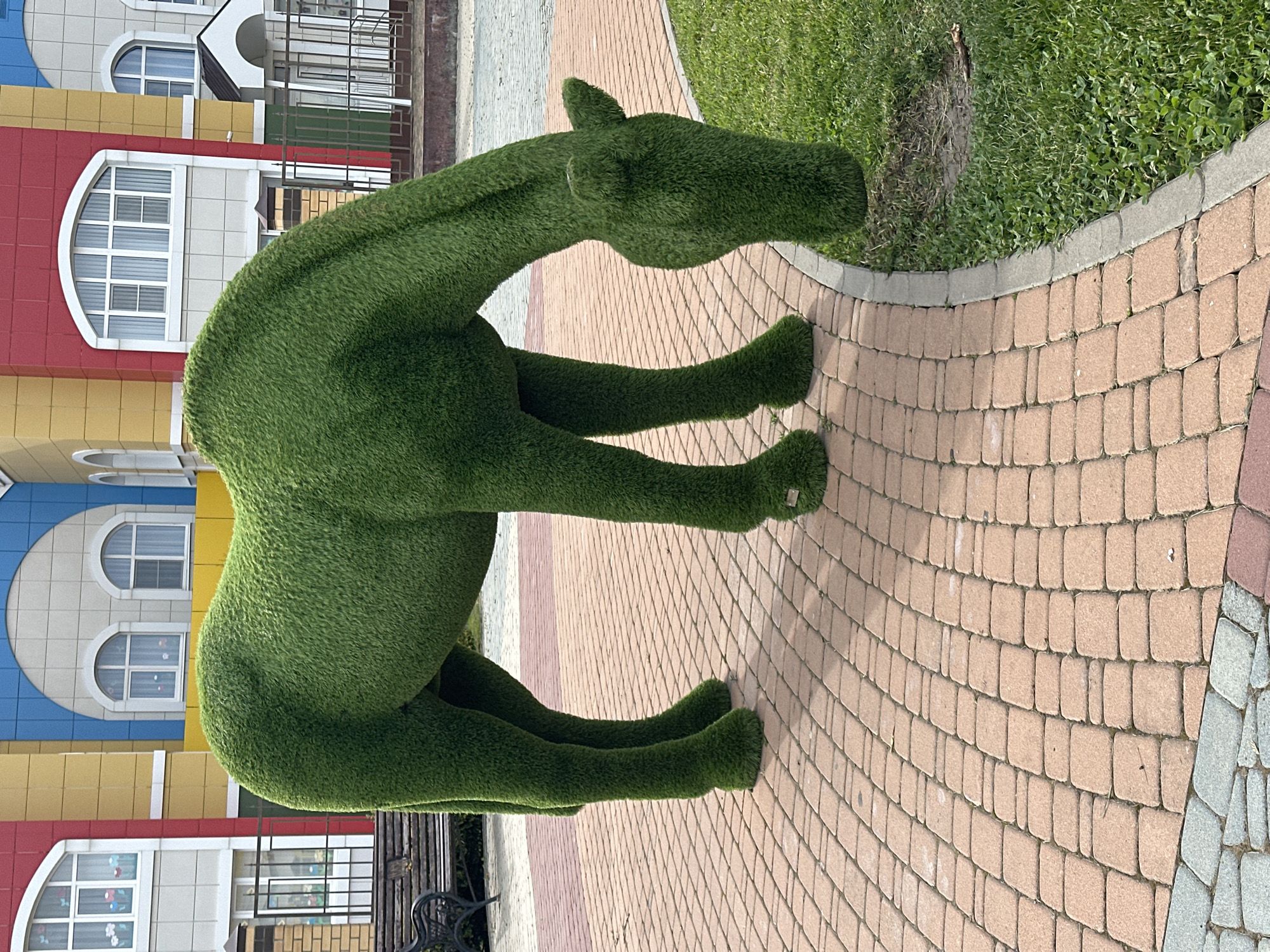 A vibrant, artificial green horse sculpture stands prominently on a reddish-brown paved pathway. The sculpture is meticulously shaped and textured to resemble a horse with its head lowered as if grazing. Behind the horse, a colorful building features a facade with red, yellow, and blue sections, adorned with arched windows with white frames. The environment appears to be an outdoor public space, possibly a park or playground, with hints of green grass and a fence in the background.