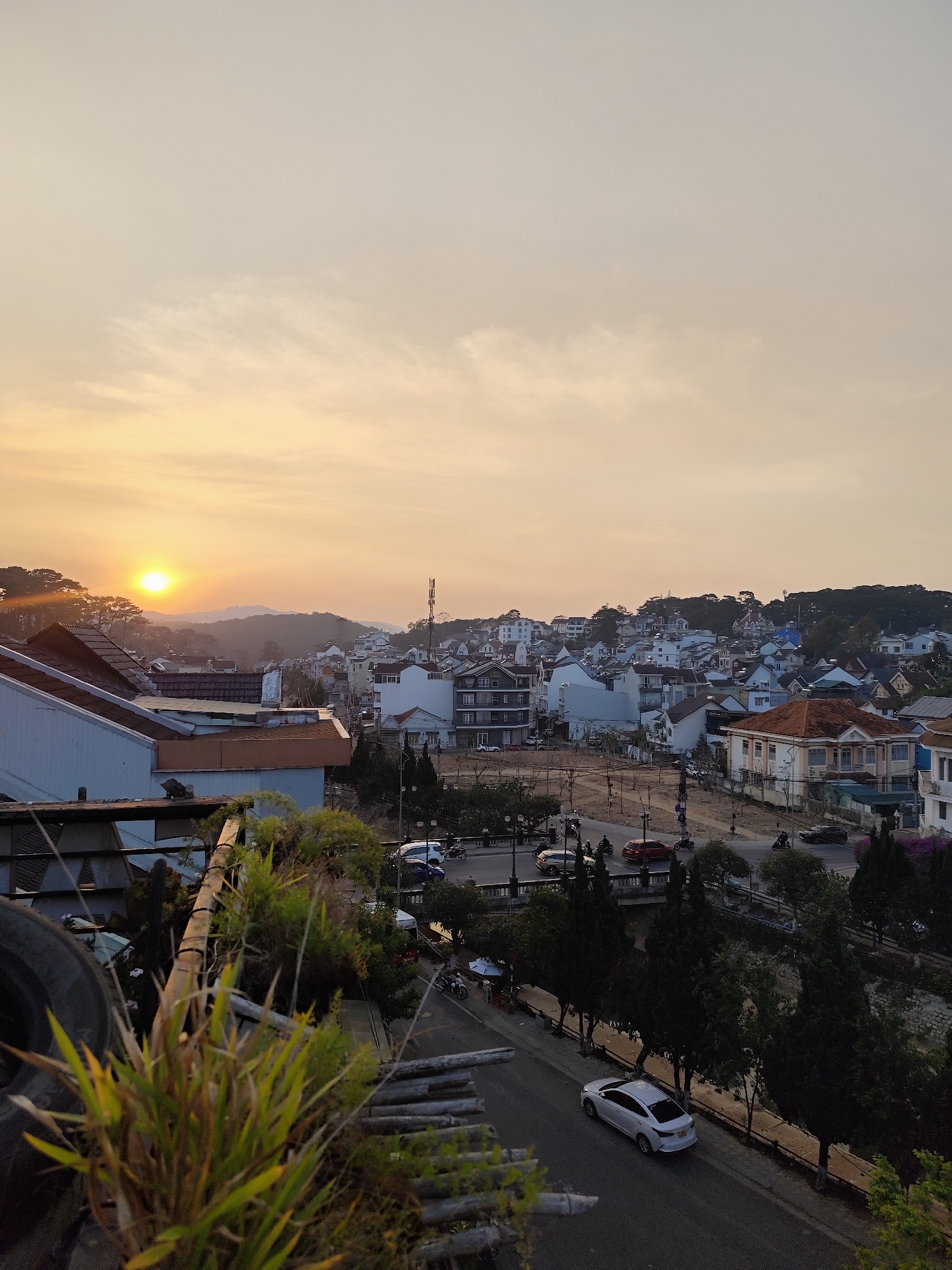 This image presents an elevated, wide-angle view of a residential and urban area in Da Lat, Vietnam, at sunset. The sky, occupying the upper two-thirds of the frame, transitions from a pale grey-blue at the top to a warm orange-yellow gradient near the horizon, with scattered, diffuse clouds. The sun, a bright, distinct orb, is positioned low on the left horizon, partially obscured by distant, forested hills.
The mid-ground reveals a dense urban landscape characterized by numerous multi-story buildings, primarily residential, with varying architectural styles, light-colored facades, and a mix of tiled and flat roofs. A tall communication tower is visible on a prominent hill in the far right mid-ground.
Below the cluster of buildings, a network of roads winds through the city. A main road occupies the lower mid-ground, where a white four-door sedan is prominently visible, parked or moving slowly on the left side of the road. Additional vehicles, including cars (e.g., a red sedan, a dark SUV) and multiple motorcycles, are observed on various streets and at intersections. A bridge-like structure is discernable crossing a narrow, lower-lying area. Extensive tree cover is integrated throughout the cityscape, lining roads and dotting hillsides.
The immediate foreground, out of focus, displays green foliage, including tall grasses, and a portion of a dark, textured, circular object, consistent with a tire, positioned on the left, suggesting the photograph was taken from an elevated structure such as a balcony or rooftop. No distinct human figures or specific interactions are clearly identifiable due to the distance.