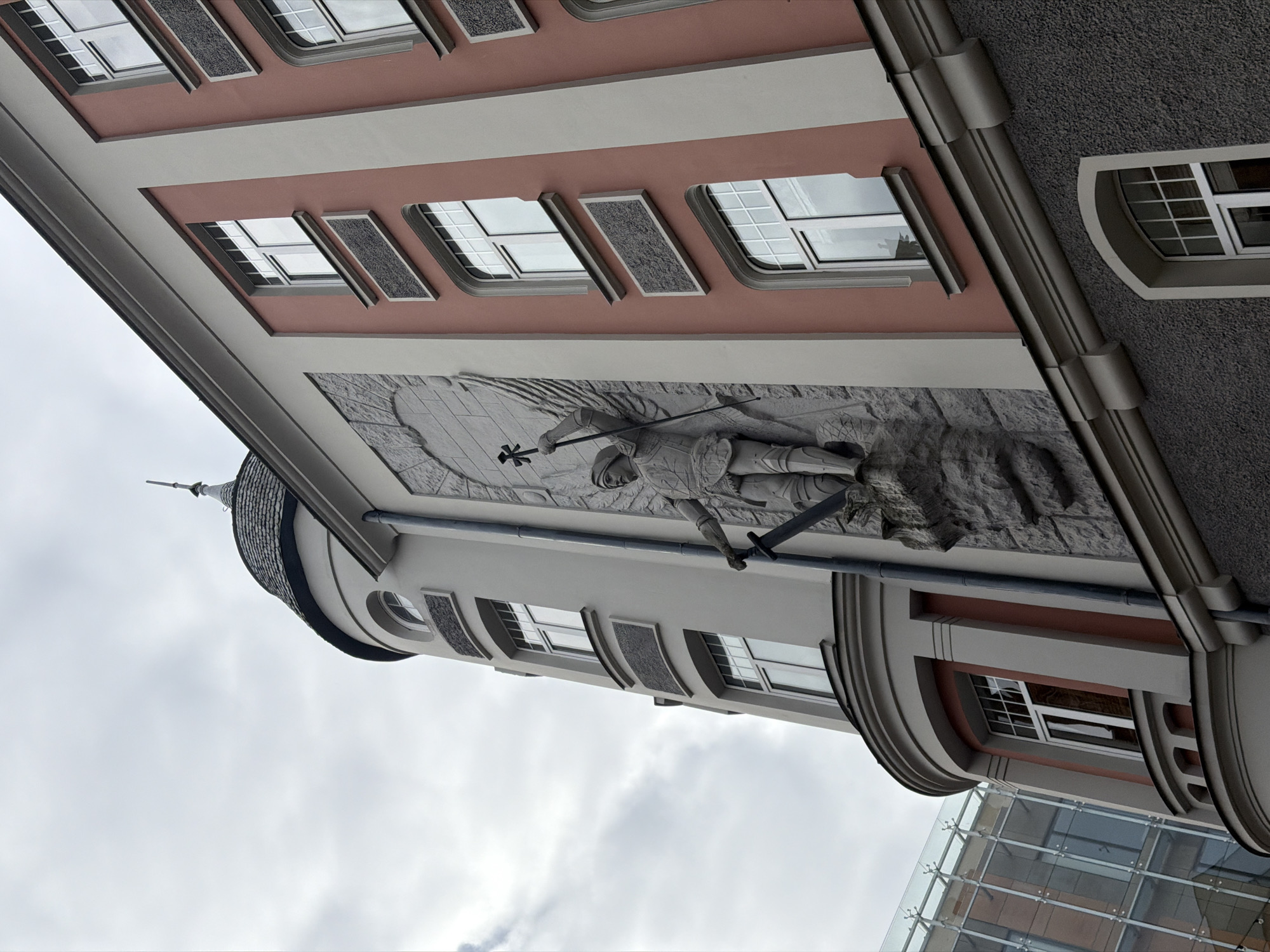 This image captures a detail of a multi-story building in Riga, Latvia, under an overcast sky. The building features a distinctive architectural style, with a prominent reddish-pink facade on its upper section. This facade is adorned with several rectangular windows, framed in grey, and flanked by textured grey decorative panels.
Below this, a large and detailed bas-relief sculpture is carved into the building's wall. It depicts a knight-like figure, heavily armored, standing in a heroic or solemn pose. The figure holds a staff, possibly a scepter or cross, in one hand and a sword, pointing downwards, in the other. Behind the knight, rays of light emanate from a textured, rocky background, giving the impression of a significant event or sacred narrative.
To the left, a small, dark-shingled conical roof of a turret or corner tower rises against the sky. The lower parts of the building show a lighter-colored facade with additional windows, some arched. In the bottom right corner, a modern glass-fronted building with a metal framework is partially visible, reflecting the cloudy sky and offering a stark contrast to the historic architecture. The time of day is daytime, but the weather is uniformly cloudy. No visible text is present in the image.