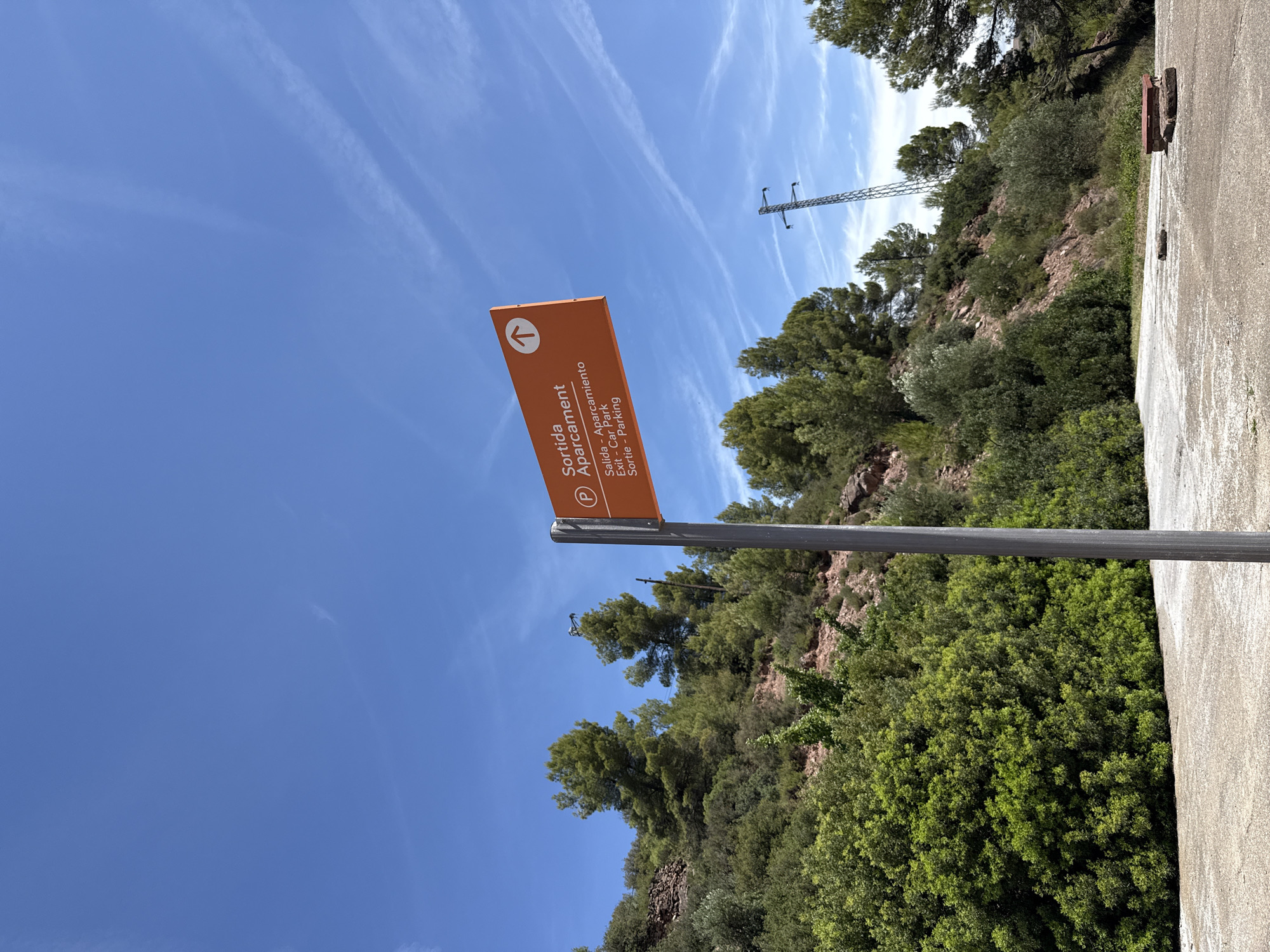 The image shows an orange directional sign indicating "Sortida - Aparcament" with translations in Catalan, Spanish, English, and French, pointing upwards with an arrow. The sign is mounted on a grey pole. The background consists of a rocky hillside covered with lush green trees and vegetation. A clear blue sky with wispy white clouds stretches above. In the distance, a tall electricity pylon is visible on the right side of the frame. The foreground is a paved area, possibly a parking lot or road, with some stones and a red object on the ground. This scene is located in the vicinity of Cardona, Spain, likely on a trail or near a viewpoint given the natural setting and signage.