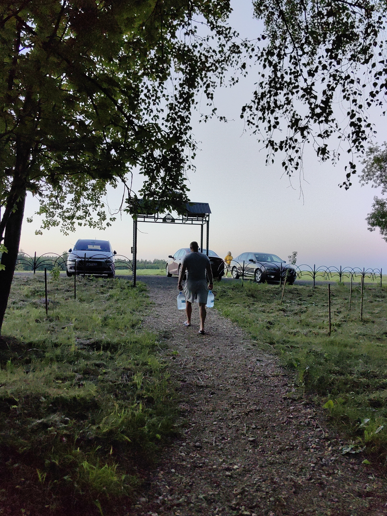 A man is walking away from the camera on a gravel path toward a covered structure. He is carrying a large clear plastic water jug in his right hand. He is wearing a dark t-shirt and light-colored shorts. In the background, there are several cars parked on the other side of a decorative fence. The scene appears to be outdoors, with grass and trees present. The sky is overcast, suggesting it might be evening or a cloudy day. The location is identified as Дзямідавіцкі сельскі Савет, Belarus.