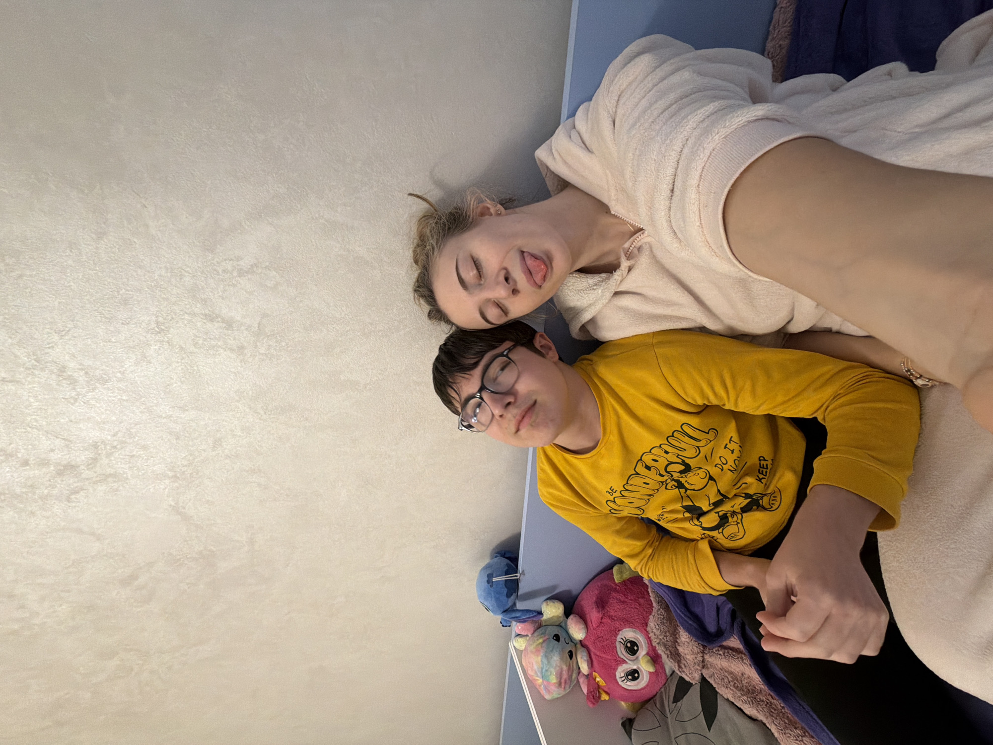 The image displays two individuals seated side-by-side in an indoor setting. The background consists of a light-colored, textured wall, with a light blue bed frame or headboard separating the subjects from the wall.
On the left is a young male, wearing a yellow long-sleeved t-shirt with black graphics. He has dark hair, wears eyeglasses, and is looking upwards and slightly to his right with a subtle, relaxed facial expression. His hands are clasped together in his lap.
On the right is a young female, dressed in a light pink, plush hooded robe. Her light-colored hair is loosely styled. Her eyes are closed, and she is sticking her tongue out playfully. Her right arm is extended downwards and slightly towards the viewer, indicating she is holding the camera for a selfie.
Behind the male, nestled against the blue frame, are several plush toys, including a large pink owl, a smaller rainbow-colored animal, and a blue creature.
The specific location within Plovdiv, Bulgaria, cannot be determined from the visual information presented in the image.