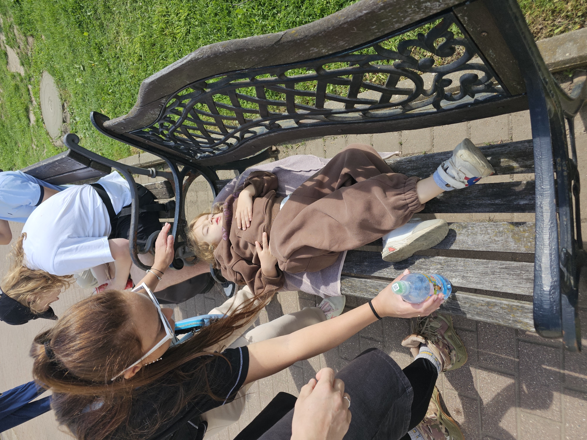 A child is lying on a bench in Borodino, Russia, attended by two adults. The child, wearing a brown outfit and an orthopedic shoe on one foot, is prone on the wooden slats of a park bench. One adult is kneeling by the child's head, while the other, wearing white-framed sunglasses, holds a water bottle. Green grass is visible behind the bench, and the surface is paved with brick-like stones.