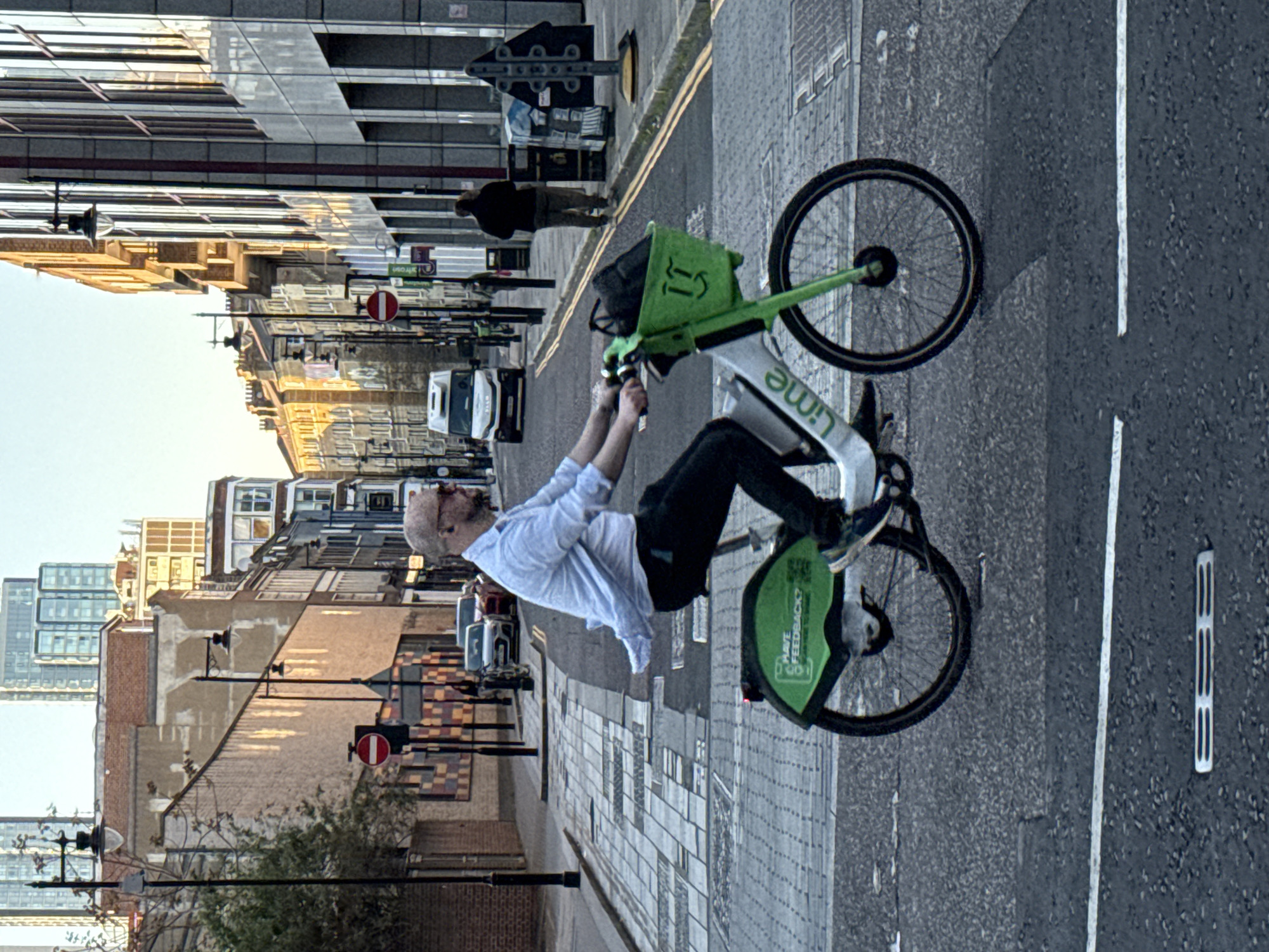 A man is riding a Lime bike on a street in London. The bike is green and white, and the man is wearing a white shirt, dark trousers and dark trainers. The street is lined with buildings, with a mix of old and modern architecture. There are street signs and lampposts along the street. Other pedestrians are visible. The street has white lines on the road.