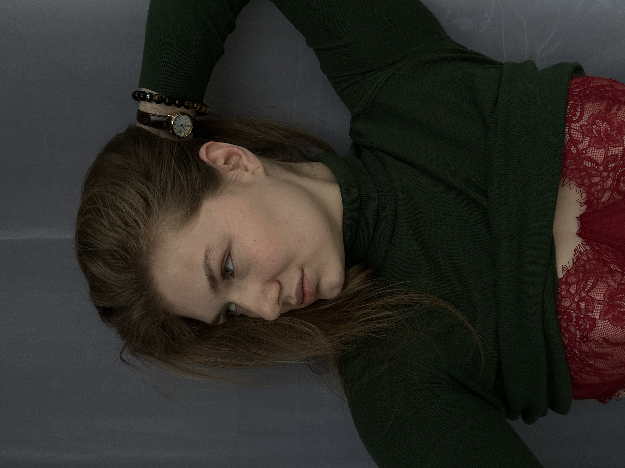A young woman is lying on a gray surface, likely a bed or a sofa. She is wearing a dark green turtleneck sweater and a red lace top is visible underneath. The woman has light skin and brown hair. She is wearing a wristwatch and a bracelet on her wrist. She is looking to her left, her expression pensive. The image does not contain any information indicating the location is Kazan, Russia.