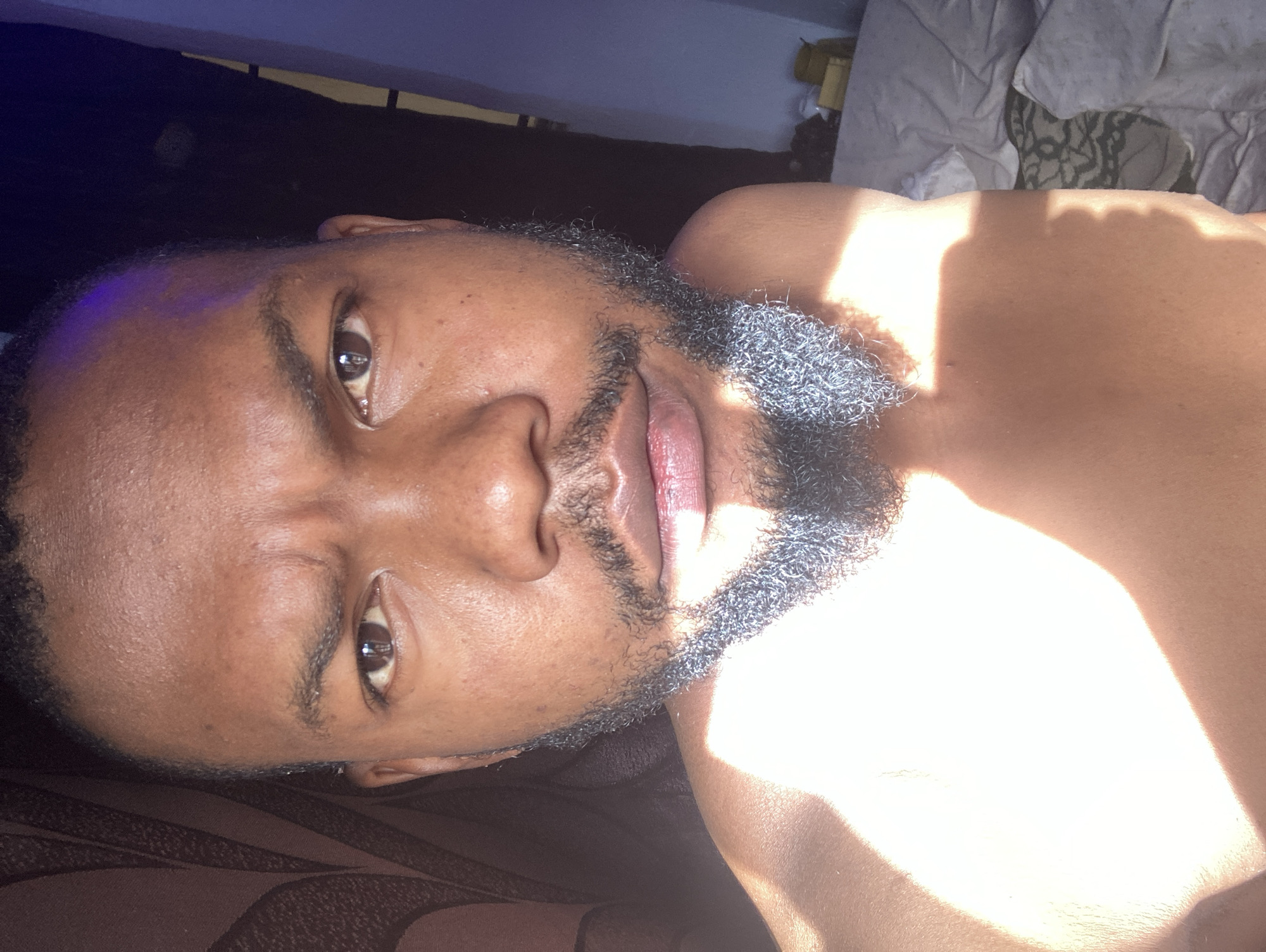 A close-up, angled portrait of a dark-skinned man with a short, dark, salt-and-pepper beard. He is lying on what appears to be a bed with brown patterned sheets. Sunlight is streaming onto his chest and the right side of his face. Behind him, there is a dark, indistinct area and part of a white and patterned pillow. There are no visible cues indicating the location of Jos, Nigeria.