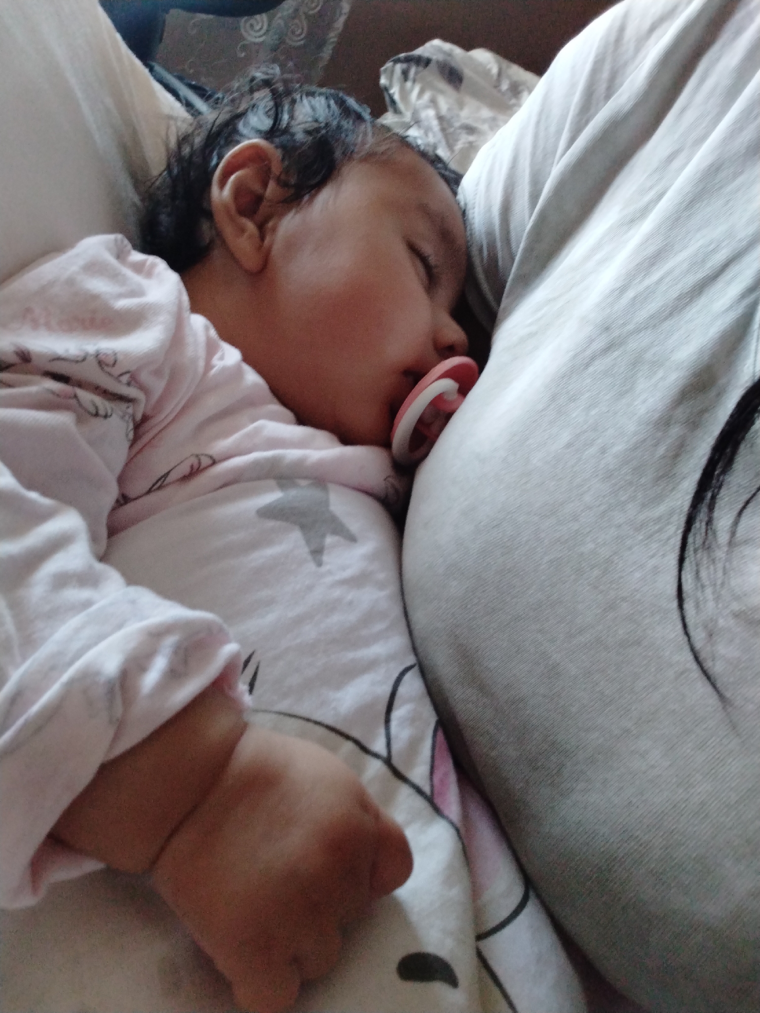 A baby sleeps peacefully with a pacifier in its mouth, nestled against a person. The baby is wearing a light pink long-sleeved shirt and is covered with a blanket featuring a star pattern. The person against whom the baby is resting is wearing a grey shirt, and strands of dark hair are visible near their shoulder. The scene is indoors. The location is Unknown, Unknown.