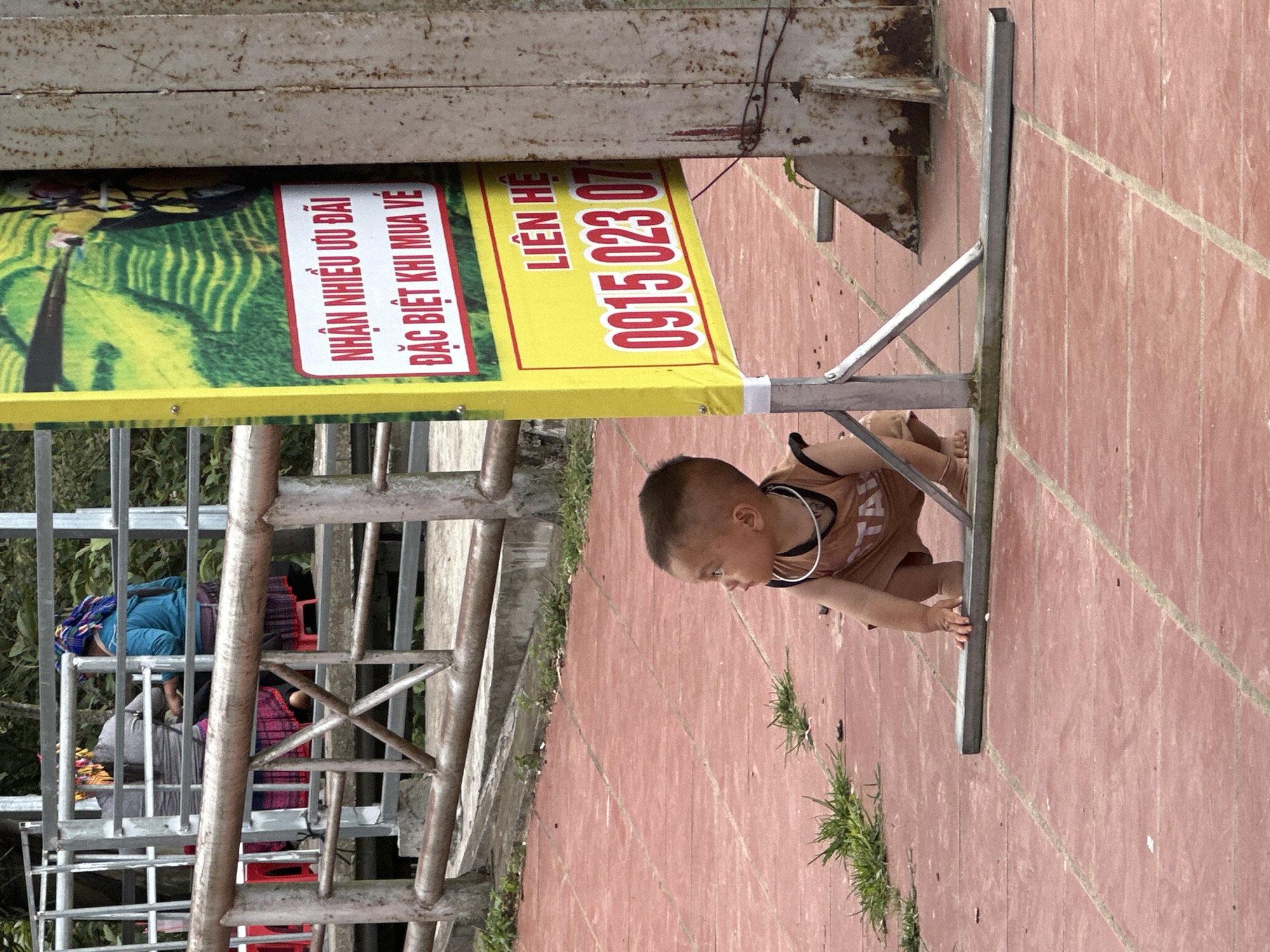 The image depicts an outdoor scene in Tu Le, Vietnam, featuring a young child in the foreground, a promotional banner, and two adults in the background.
In the foreground, a barefoot toddler, dressed in a brown sleeveless top and matching shorts, crouches on a reddish-brown tiled or paved surface. The child's hands are placed on the ground near the base of a sign. A light-colored, possibly metallic, ring is visible around the child's neck. The child gazes towards the left side of the frame. Scattered patches of green grass grow through cracks in the pavement.
A yellow vertical banner, supported by a metal frame, stands prominently in the midground. The banner displays text in red and white Vietnamese characters, with an image of green terraced fields at its top. Visible text includes "NHẬN NHIỀU ƯU ĐÃI ĐẶC BIỆT KHI MUA VÉ" (Receive many special offers when buying tickets) and "LIÊN HỆ 0915 023 07" (Contact 0915 023 07), with the last digit of the phone number obscured. To the right of the banner, a corroded, rusty metal support structure is partially visible. A brown, rusted metal railing extends horizontally across the scene, separating the foreground and midground from the background.
In the background, behind the brown railing, a more complex grey metal railing system with diagonal cross-supports is present. Two adults, likely women based on traditional attire, are partially visible behind this railing. One wears a blue long-sleeve top and a patterned head covering, while the other wears a grey top and a similar head covering. Both appear to be wearing traditional patterned skirts. Distant green foliage forms the furthest background element. The ground beneath the railings includes concrete structures. The scene is illuminated by ambient daylight.