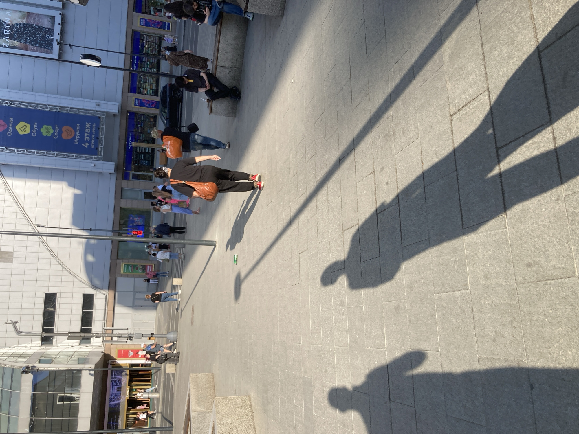 The image depicts a city scene, likely in Moscow, Russia, given the Cyrillic text on the building advertisements. The view is angled, showing a wide pedestrian area with various people moving about. Several individuals are walking, some appear to be window shopping, and others are seated.
The environment consists of modern buildings, one with large store windows, and a paved pedestrian square. The light suggests it's daytime. Shadows of objects or structures are visible across the paving stones.
Essential details include:
* People: A diverse group of pedestrians engaging in various activities.
* Objects: Street furniture, advertisements, and possibly a vehicle.
* Actions: Walking, sitting, shopping.
* Environment: Modern urban setting with paved square and large buildings.
* Signs written in Cyrillic.