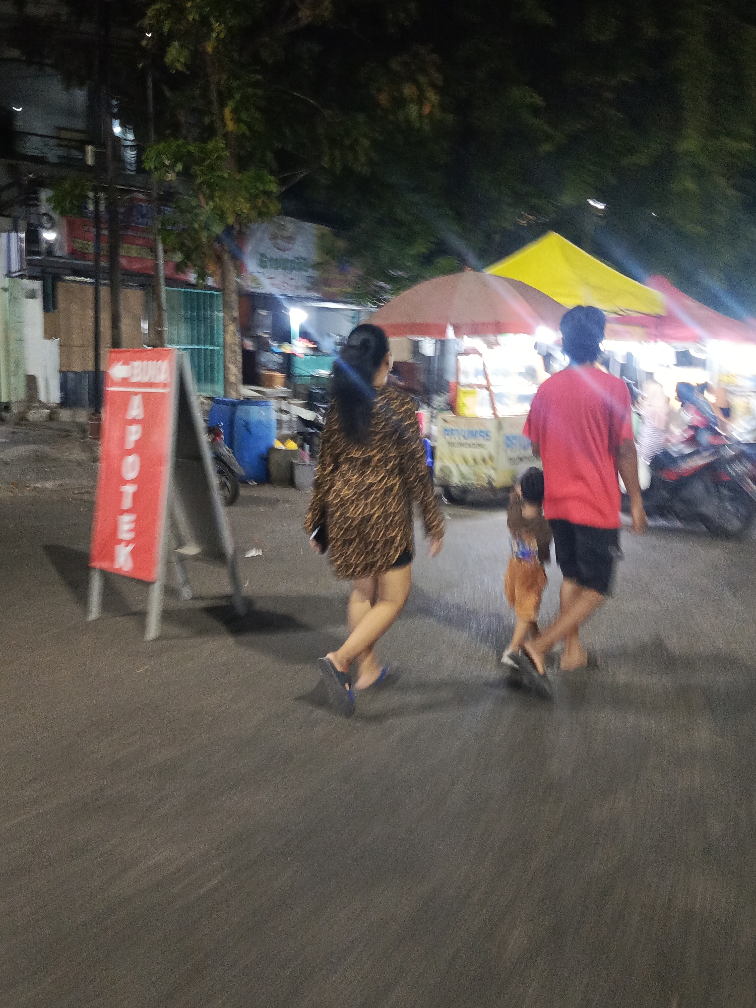 This image depicts a street scene at night in Surabaya, Indonesia, likely near a local market or food stalls. In the foreground, a family walks away from the camera. The mother, on the left, wears a leopard-print dress and blue flip-flops. She is holding a black phone in her left hand. Beside her, a man in a red t-shirt and black shorts walks with his son, who is wearing an orange shirt and pants. They appear to be moving from left to right.
To the left of the family is a red A-frame sign with white text that reads "BUKA APOTEK", indicating an open pharmacy. Behind the sign, there are some buildings and a green fence. To the right of the family, illuminated by bright lights, are market stalls with colorful awnings, one yellow and one red. A motorcycle is parked to the far right. The ground is asphalt, and there are trees and overhead wires visible in the background, suggesting an urban environment. The overall impression is of a lively evening scene in a busy part of the city.