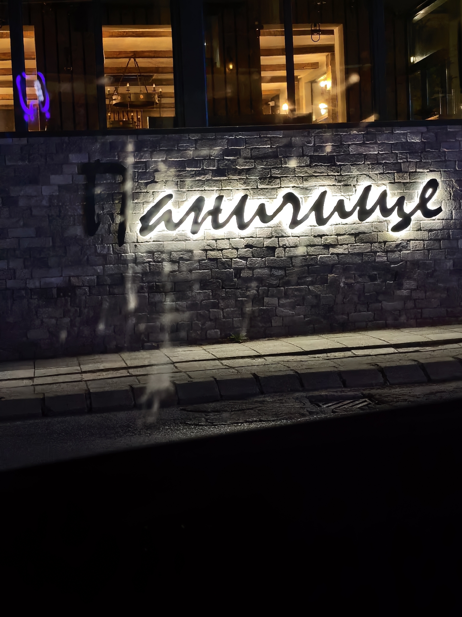 Here is a detailed description of the image:
**Location:** The image appears to be taken outdoors in an urban area, likely at night, in the city of Unknown, Unknown.
**Environment:** The setting is dark with artificial lighting highlighting a brick wall and a sidewalk. Above the brick wall is a building.
**Subjects and Objects:**
* A large sign in a cursive style is mounted on the brick wall. The sign is backlit, emitting a bright light.
* The wall is constructed of gray bricks.
* A sidewalk made of gray pavers is adjacent to the brick wall.
* A dark road or street is visible.
* A building is visible above the wall. The interior shows wooden beams and lighting fixtures.
* A drain is visible in the street.
**Actions/Interactions:**
* The sign appears to be advertising or labeling a business or establishment.
* The lights are illuminating the building and the sign.
**People:**
* No people are visible in the image.
**Overall Impression:** The image presents an exterior view of a building or establishment at night, focusing on the illuminated signage.