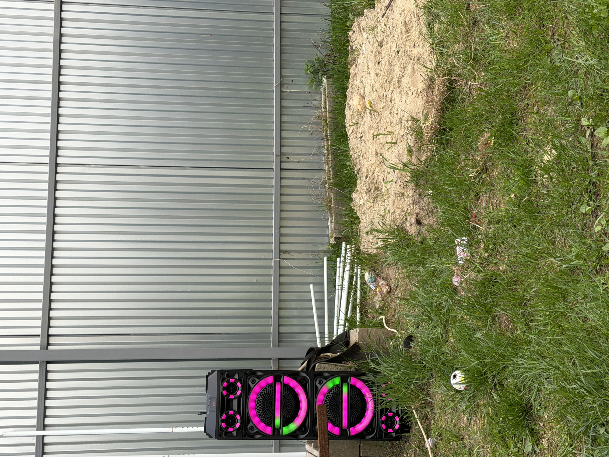 The image shows an outdoor scene with a corrugated metal fence in the background. To the left of the fence, there's a large black speaker with circular LED lights in pink and green. Some white PVC pipes are lying near the speaker and the fence. To the right, there is a mound of dirt and dense green grass. The location is likely Sverdlovskiy, Russia based on the user’s request. There are no people visible in the image. The overall impression is of an unkempt outdoor space next to a structure with a metal fence.