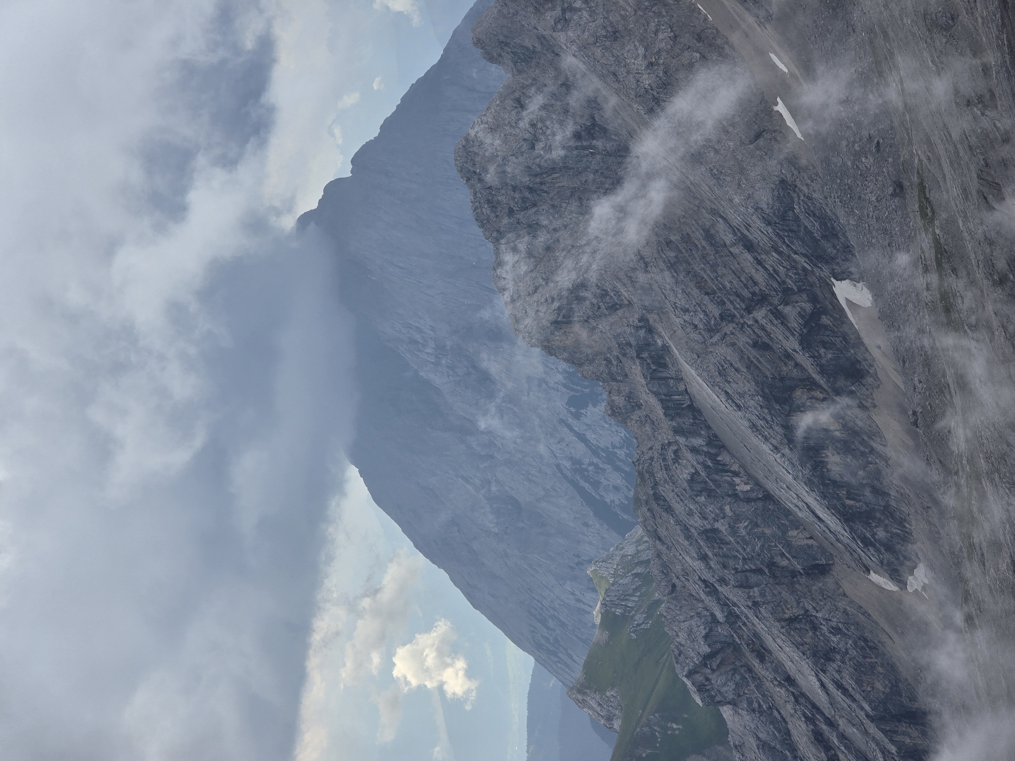 The image shows a view of the Zugspitze mountain range in Garmisch-Partenkirchen, Germany. In the foreground, there are rugged, gray, rocky mountain slopes with patches of snow and low-lying clouds or fog swirling around them. In the middle ground, another rocky peak rises, also streaked with rock and some sparse vegetation. In the background, a much larger, imposing mountain peak dominates the scene, partially shrouded in dark, heavy clouds. The sky is overcast with varying shades of gray clouds, with a small, white cumulus cloud visible to the left of the distant peak. The overall impression is a dramatic, high-alpine landscape under a moody sky.
