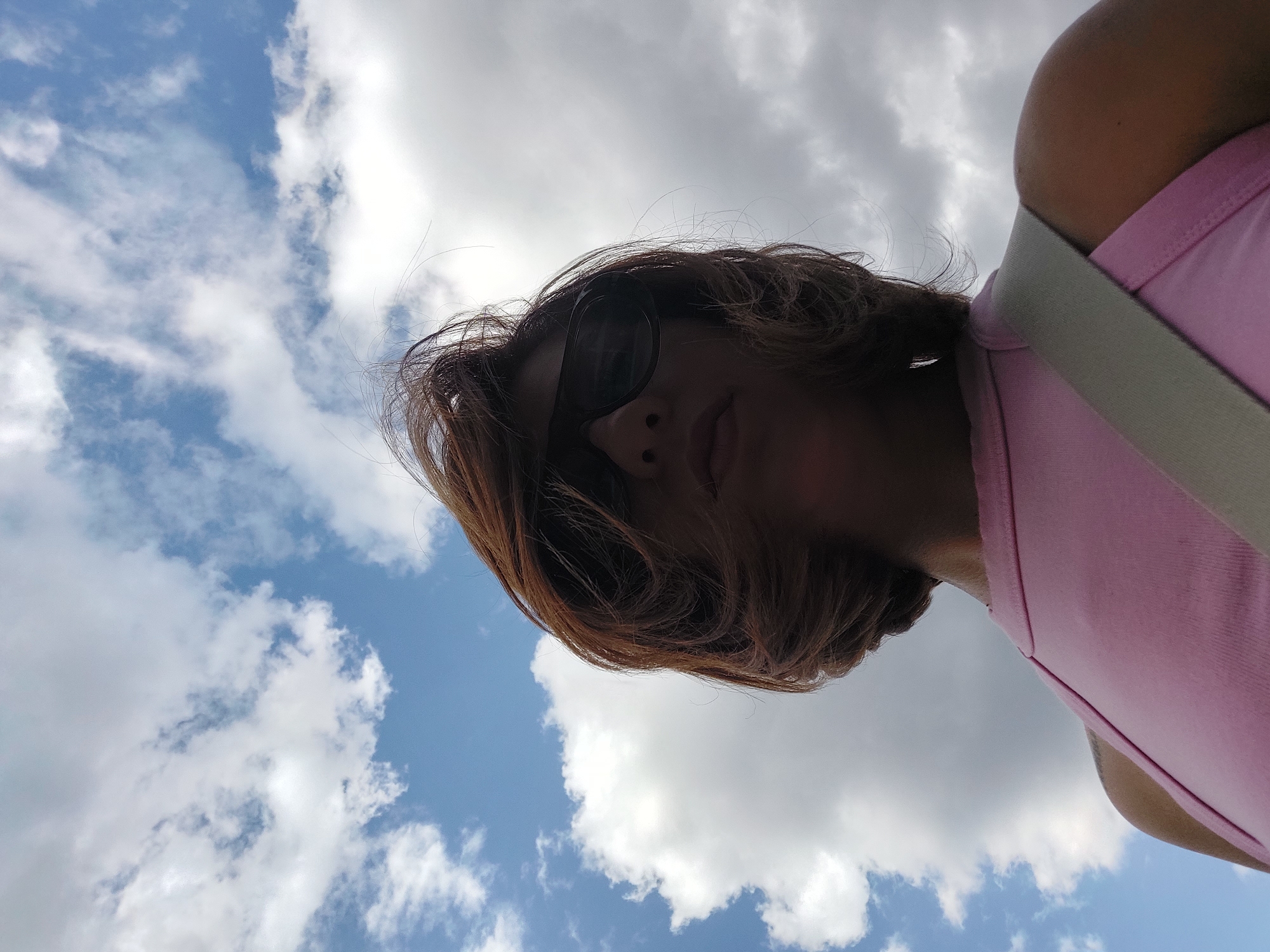 A low-angle, wide shot captures a person from the chest up, looking upwards at a partly cloudy sky. The person, wearing a pink top and dark sunglasses, has shoulder-length brown hair that is windswept. The background is dominated by a bright blue sky with scattered white clouds. A light-colored strap crosses diagonally across the person's chest. The image was taken outdoors on a bright, possibly windy day. The location is not specified.