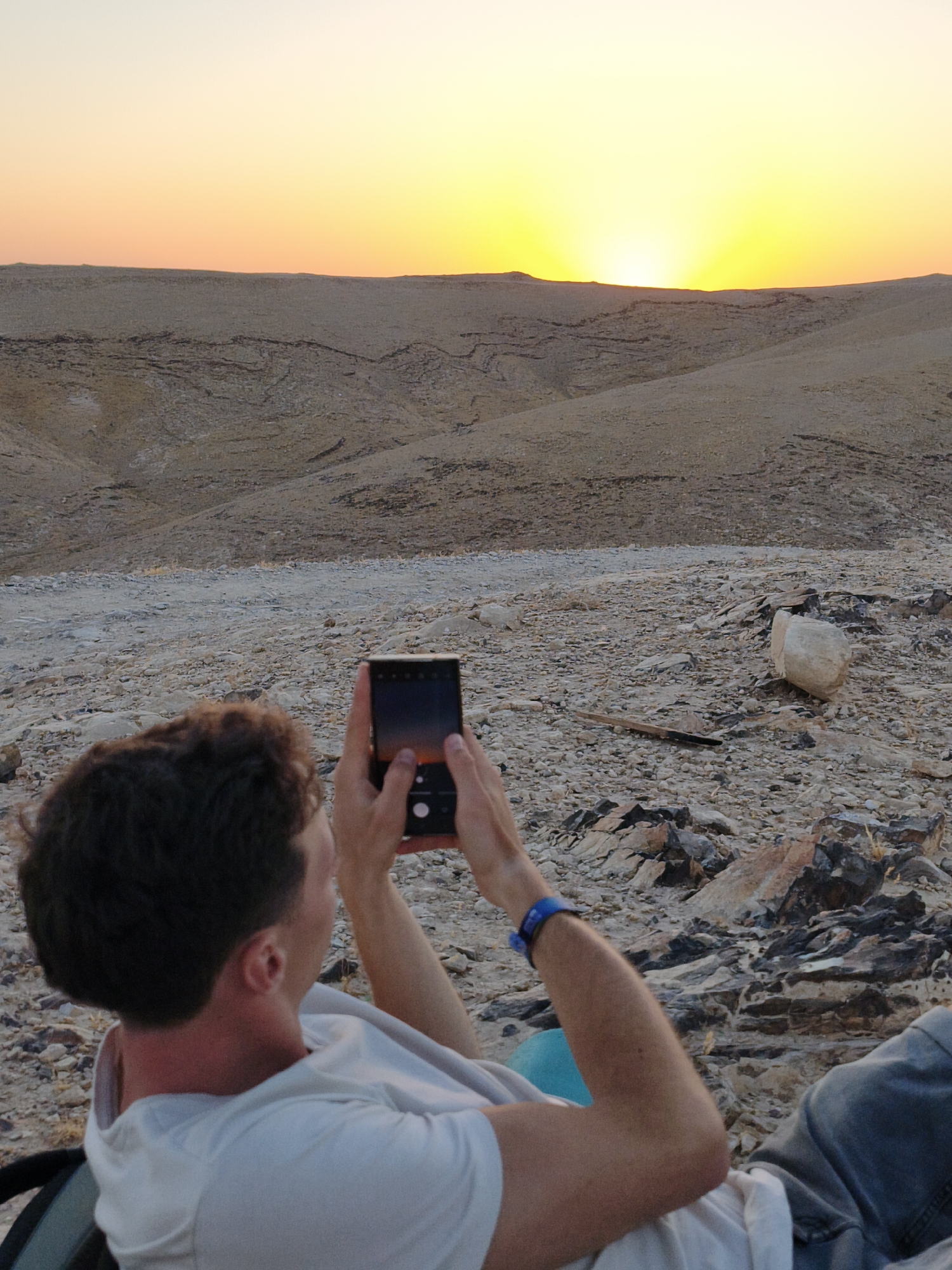 A person is shown from behind, lying down on a rocky desert terrain, holding a smartphone and taking a photograph of the sunset. The person is wearing a white t-shirt and blue jeans, and a blue wristband is visible on their left wrist. The terrain is arid and sparsely vegetated, with hills and valleys stretching into the distance. The sky is a gradient of orange and yellow, with the sun setting on the horizon. The scene appears to be located in the city of Keidar, Palestinian Territories, a region known for its desert landscapes.