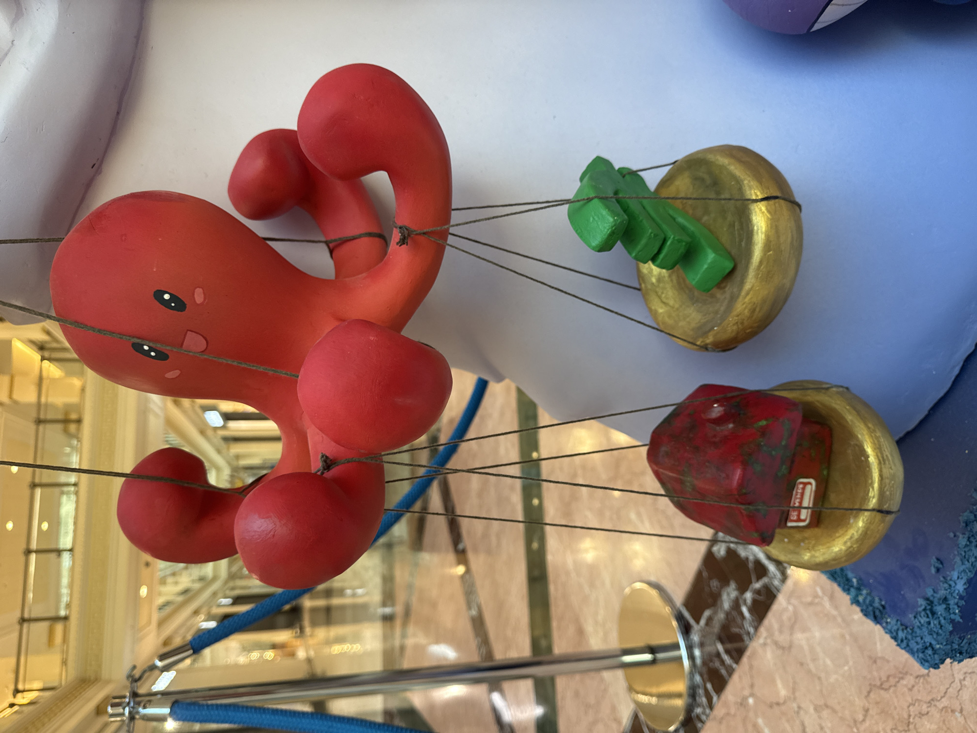 A close-up, medium shot captures a red octopus decoration in an indoor setting. The octopus, with wide eyes and a smile, is suspended by strings. From two of its tentacles, smaller objects are hung. On the left, a golden dish holds a red, gem-like object with a small white label that reads "SE VENDE." On the right, a similar golden dish supports a stack of three green rectangular blocks. The background shows a blurred interior with marble flooring and what appears to be a shopping mall or hotel lobby, indicated by stanchions with blue ropes and railings.