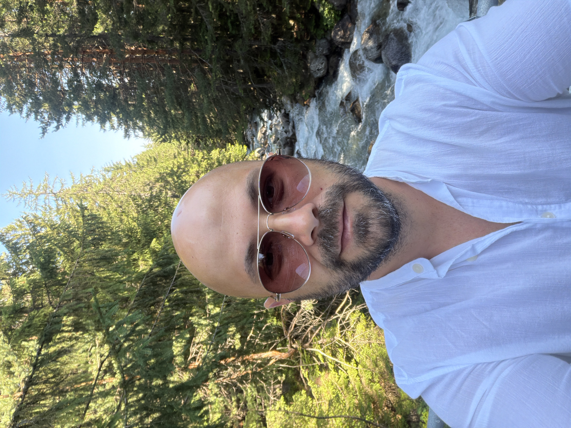 A man is taking a selfie in the mountains. He is bald, has a beard, and is wearing sunglasses and a white shirt. He is standing in front of a stream with trees on either side. The image appears to be taken during the daytime, with the sun shining. The location is the Pirin National Park, near Bansko, Bulgaria.