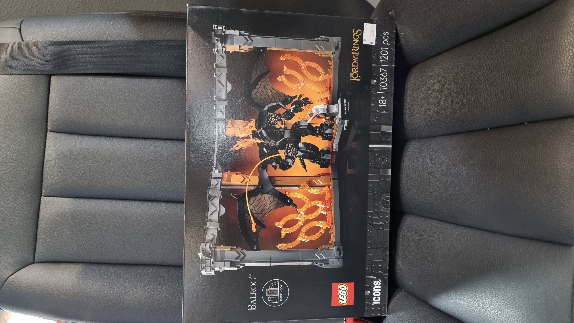 A new, unopened LEGO set box, specifically "BALROG™" from "THE LORD OF THE RINGS™" (Set 10367, 1201 pieces, aged 18+), rests on a dark grey or black textured car seat. The box's front art depicts a LEGO rendition of the Balrog, a large winged, flaming creature, confronting a Gandalf minifigure holding a sword on a narrow bridge, with the inscription "You shall not pass!". An emblem indicates the set is designed as a "BOOK NOOK". A white sticker on the bottom right of the box shows a price of €132.89, with a partially obscured higher price (e.g., €2xx.90) crossed out above it. The car seat features vertical pleats, and a black seatbelt is visible on the right side of the seat's backrest. The scene is located in the city of Sofia, Bulgaria.