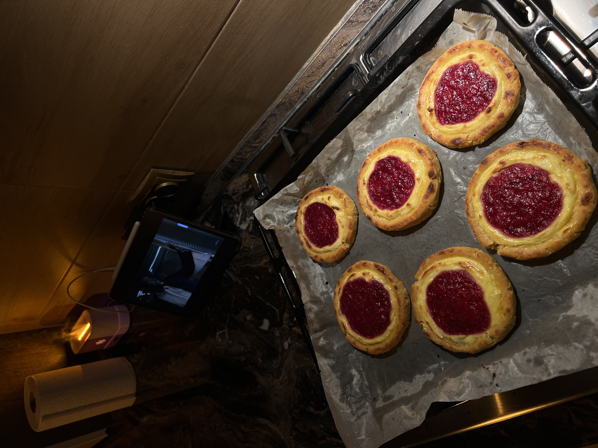 The image displays a tray of freshly baked pastries with a red jam filling, resting on a sheet of parchment paper. The pastries are arranged on a dark countertop that appears to be a kitchen surface, likely in an apartment or house. In the background, a roll of paper towels and an electronic device are visible, with ambient lighting suggesting an indoor setting. The context does not provide any information about the location in the city of Ternopil, Ukraine, or any specific details about people, actions, or interactions.