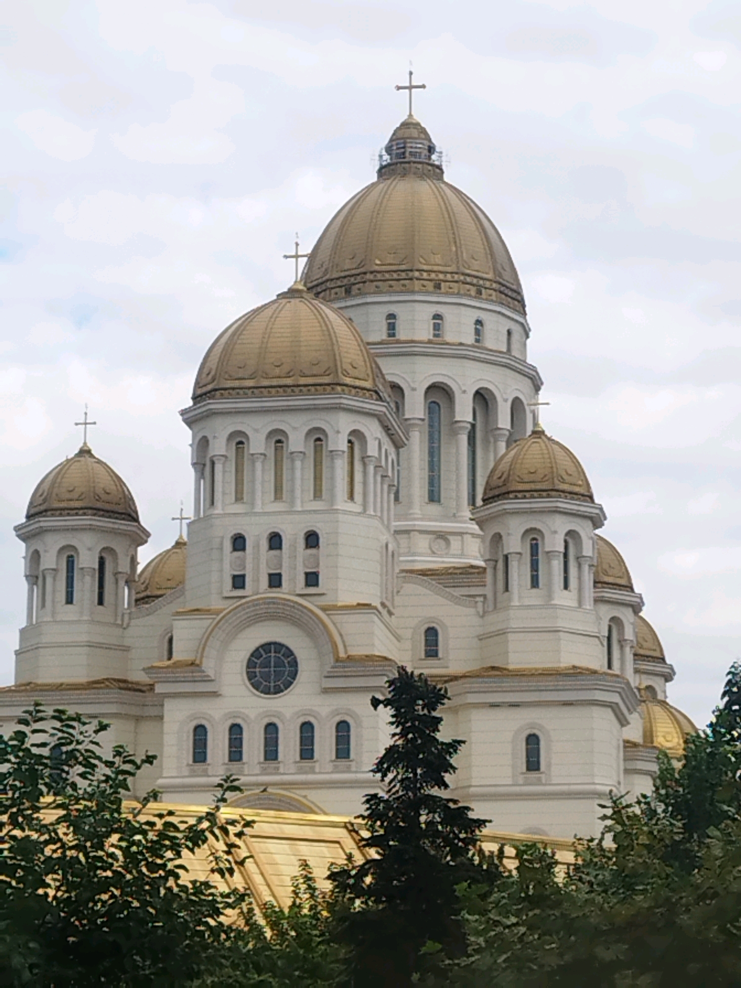 The image shows a close-up, frontal view of the Romanian Orthodox Patriarchal Cathedral, located in Bucharest, Romania. The cathedral is a large, white building with multiple golden domes, each topped with a cross. The architecture features arches, columns, and tall, narrow windows. Some of the smaller domes are visible on the sides and front of the main structure. In the foreground, lush green trees and foliage partially obscure the lower part of the cathedral, with the golden roofs of some sections of the building also visible peeking through the leaves. The sky above is overcast with soft, diffused light. The scene is static and depicts the exterior of the religious structure.