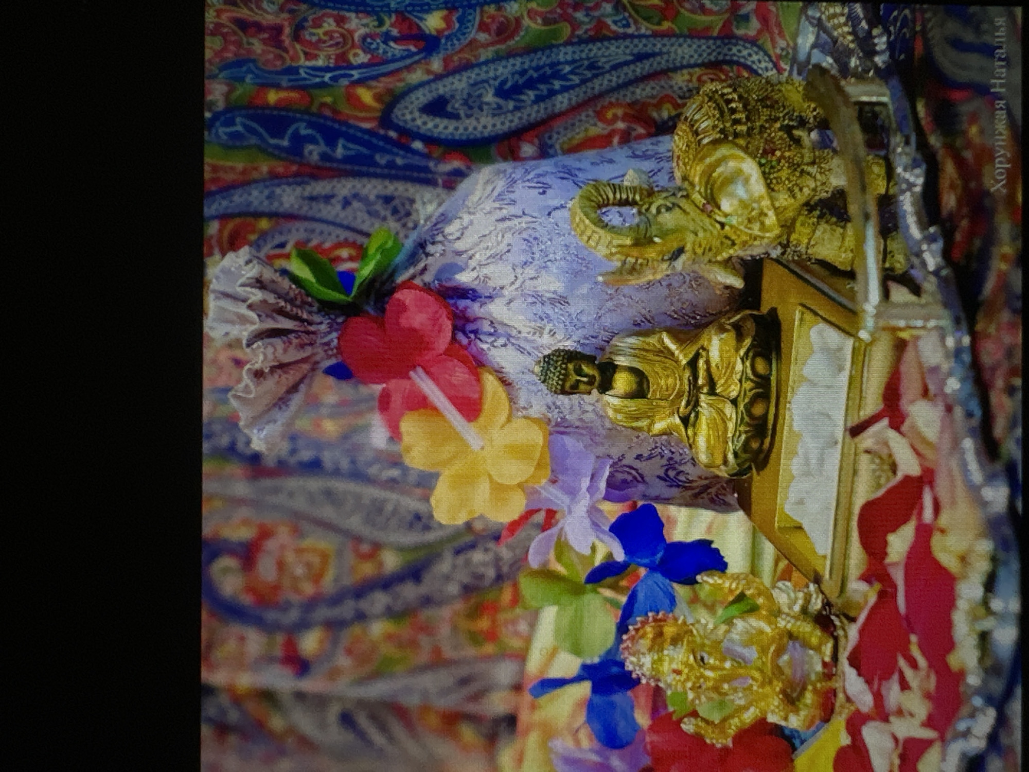 The image displays a collection of religious figurines and decorative items, likely arranged for an altar or display. In the foreground, a golden statue of Ganesha, the Hindu deity of wisdom and prosperity, is prominently featured. To the right of Ganesha, a golden statue of a seated Buddha is visible. Adjacent to the Buddha, a golden elephant figurine with intricate detailing stands. These figurines are placed on a golden altar. Surrounding the altar are various decorative elements, including colorful artificial flowers in shades of yellow, red, blue, and purple, and what appear to be rose petals scattered across the surface. A light purple, fabric-covered object, possibly a pouch or bag with decorative pleats, is situated behind the figurines, adorned with more artificial flowers. The background is a colorful, patterned fabric, possibly a tapestry or shawl, with paisley-like motifs in shades of blue, red, and yellow. The overall composition suggests a devotional or celebratory context. The scene does not provide any specific information or visual cues that would indicate its location within the city of Luhansk, Ukraine.