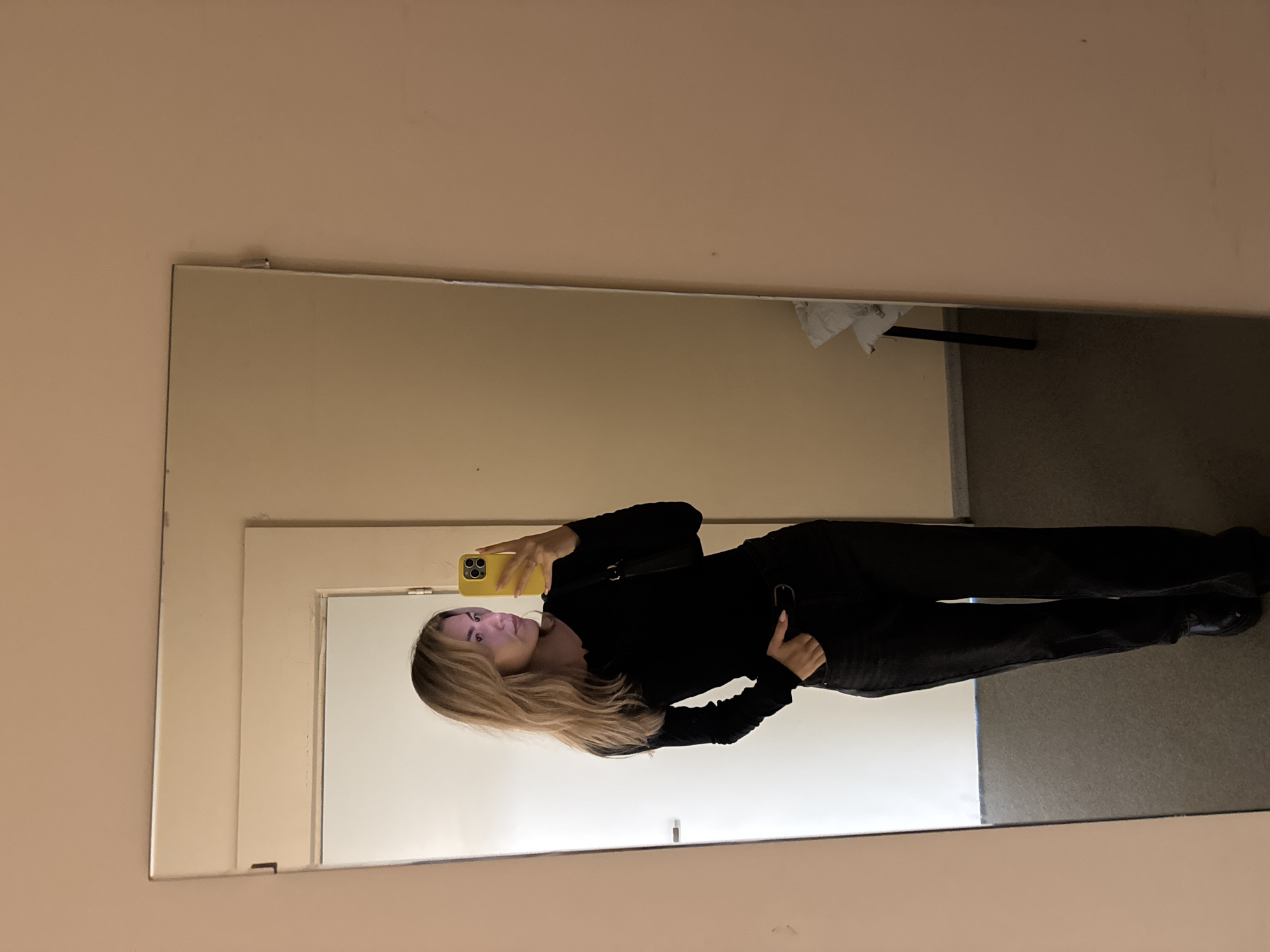 A woman is taking a full-body mirror selfie in an unidentified room. The mirror reflects a neutral-colored wall, a doorway, and what appears to be a gray carpeted area. The woman is wearing dark clothing and holding a yellow smartphone. Her hair is light brown with blonde highlights. The location is listed as Unknown, Unknown.