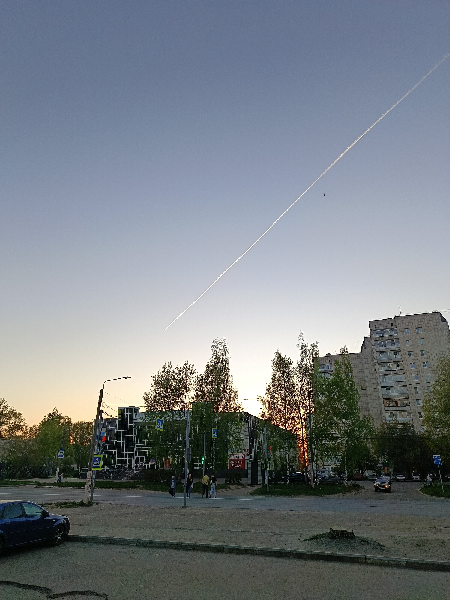 The image depicts a street scene in Chaikovsky, Russia, at dusk or dawn. The sky is light blue, transitioning to orange near the horizon. A long contrail stretches across the sky, with a small dark object near it. In the foreground, a blue car is parked on the left side of the frame. A crosswalk leads across the street, where a group of pedestrians is walking. Behind the pedestrians is a building with a glass facade and multiple pedestrian crossing signs. To the right of the glass building, trees and a multi-story apartment building can be seen. A car is driving along the street on the right. The ground is a mix of pavement and patches of grass. A tree stump is in the very foreground.
