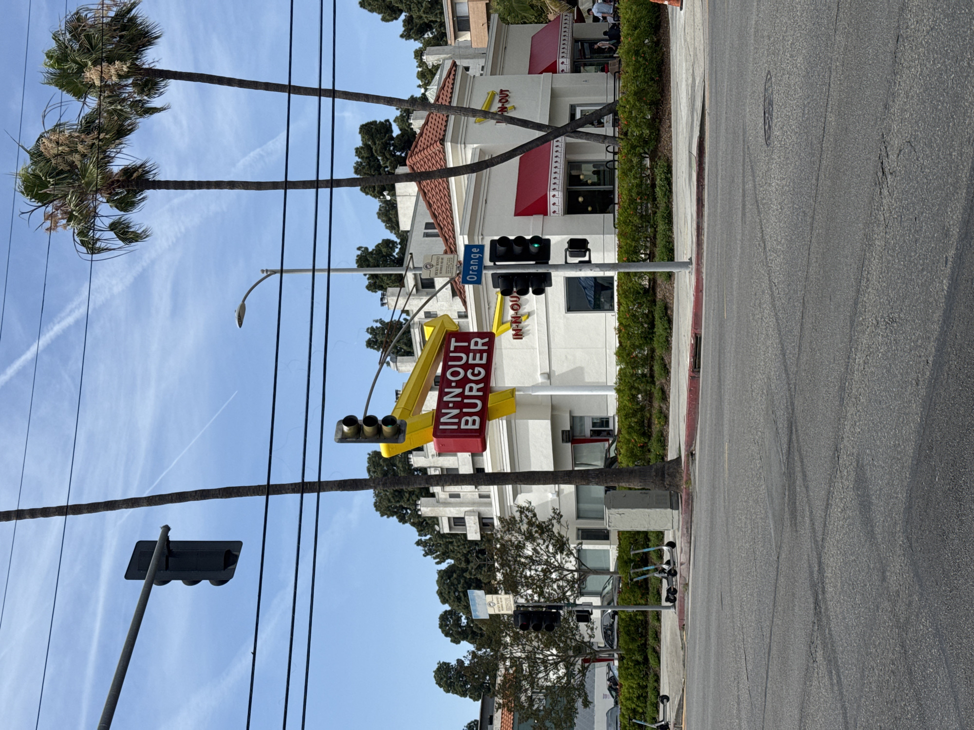 The image captures a vibrant daytime street scene in what appears to be Los Angeles, California. The main subject is an iconic In-N-Out Burger restaurant, prominent on the right side of the street. The building is white with red tile roofing and red awnings, displaying the "IN-N-OUT" logo in yellow on its facade. A large, distinctive red and yellow sign with "IN-N-OUT BURGER" written vertically stands tall at the corner of the property, partially obscured by power lines and a traffic light pole.
In the foreground, a paved street stretches across the bottom of the frame, showing tire marks and a manhole cover. A sidewalk with a hedge runs alongside the street, where several electric scooters are parked. Two tall palm trees dominate the left side of the image, their fronds reaching into a clear, bright blue sky adorned with wispy white clouds and a faint airplane contrail. Numerous power lines crisscross the sky, adding to the urban feel.
Traffic lights are visible, with one set showing a green light. A blue street sign on a pole clearly reads "Orange", indicating "Orange Street". Another sign or plaque, partially legible, states "MICKEY ROONEY WALK OF FAME", a strong indication that the location is in Hollywood, Los Angeles. The time of day appears to be bright and sunny, likely morning or afternoon, with no discernible human activity or event taking place beyond the general hustle of a city street. A few people are faintly visible near the restaurant's entrance and in the background, but no specific individuals are clearly identifiable.