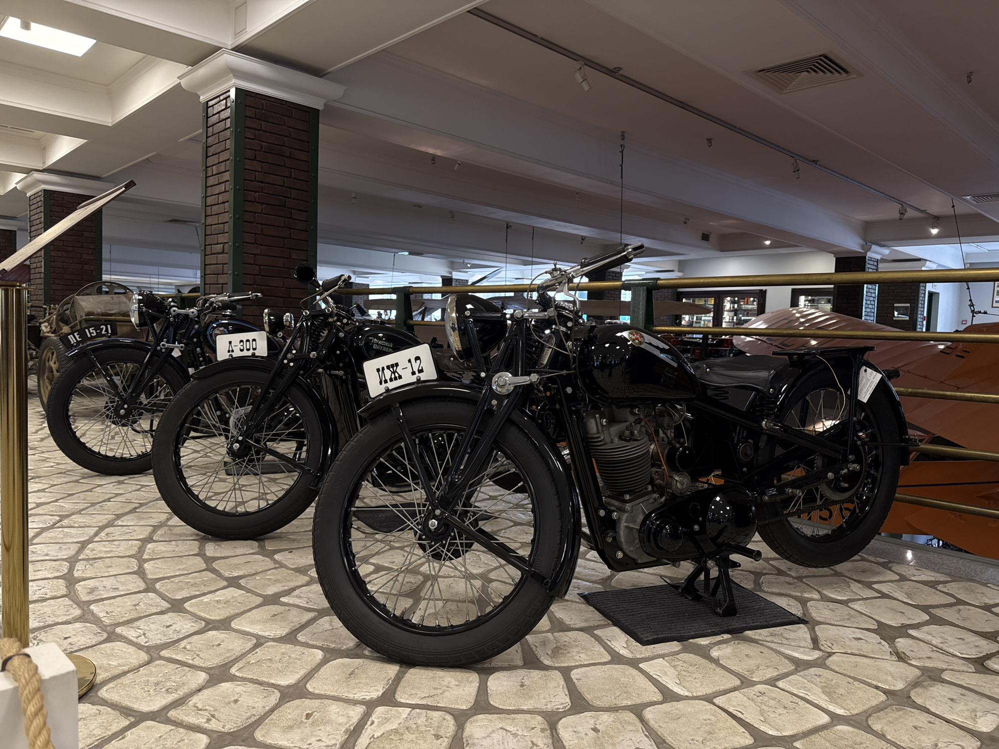 The image shows a display of several vintage black motorcycles parked on a stone floor inside a museum. The motorcycles are positioned in a row, with the closest one in the foreground to the right. The closest motorcycle is detailed, showing its engine, black seat, spoked wheels, and handlebars. A license plate on this motorcycle reads "MX-12". To the left of this motorcycle, other similar vintage motorcycles are visible, some with license plates reading "A-300" and "ME 15-21". The background reveals a museum setting with brick columns, exposed ceiling beams with track lighting, and a display case or exhibit in the distance. The environment appears to be well-lit and exhibits a sense of historical preservation. The location is the estate museum-Arkhangelskoye in Moscow Oblast, Russia.