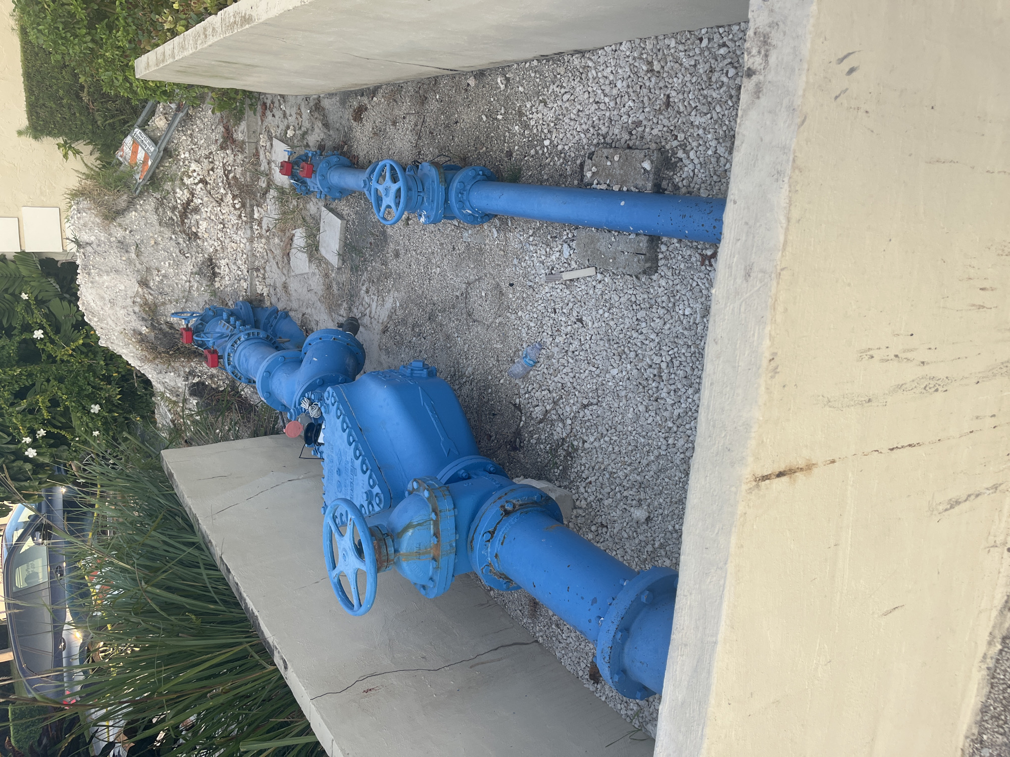 In an outdoor setting, two large blue pipes with red valves and wheel-shaped handles are visible. The pipes are set in gravel and are surrounded by concrete structures. The larger pipe, in the foreground, is thick and appears to be a main water line. It has a large, blue flow meter attached to it. The smaller pipe, further back, is also blue and has similar valves. In the background, there is a pile of white rocks and some greenery, including tall grass and bushes. A car and a road sign are also visible in the background, suggesting an urban or suburban environment. The scene appears to be a utility infrastructure setup, possibly for water management or distribution. The lighting suggests it is daytime, with some shadows indicating the sun's position.