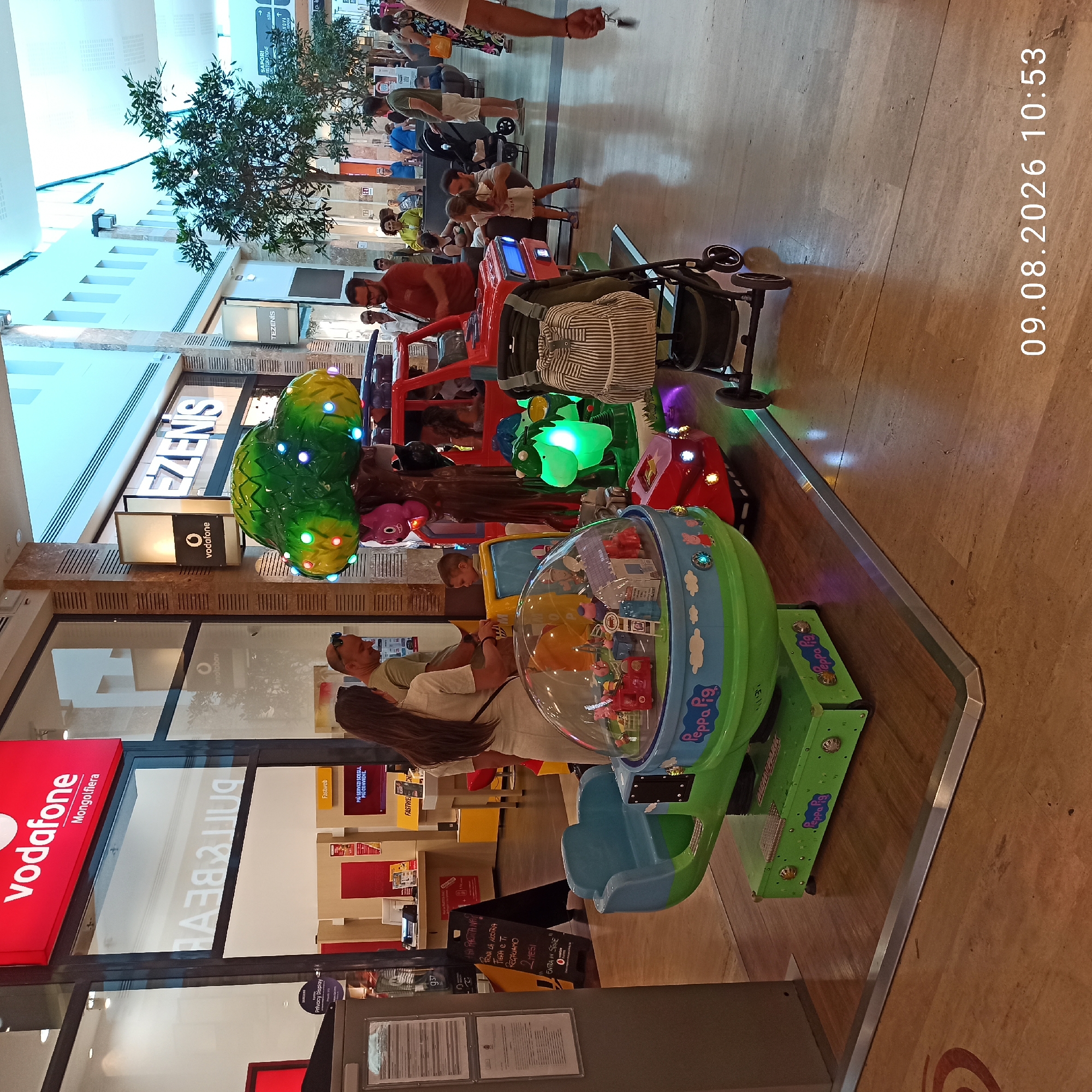 The image shows an indoor shopping center, likely in Molfetta, Italy, as indicated by the "Vodafone Mongolfiera" signage. In the foreground, a brightly colored Peppa Pig themed children's amusement ride is visible, featuring a dome with miniature scenes and a vehicle-shaped seat. Adjacent to the ride is a stroller. A man and a woman are standing near the ride, looking at a young boy who is seated on a red toy car, another part of the amusement ride. In the background, various shops are visible, including "Vodafone" and "TEZENIS". Several other people are scattered throughout the shopping center, some walking, some standing. The floor is made of large, light-colored tiles. The overall ambiance is that of a public, commercial space during daytime.