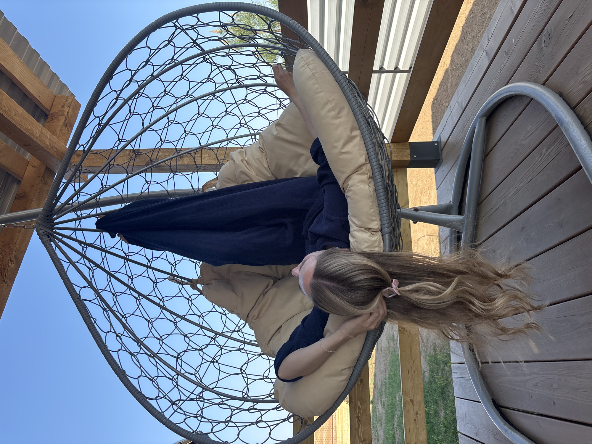 A woman is lying upside down in a hanging chair. The chair is made of a gray metal frame with a woven seat. The woman is wearing a dark blue dress, and she has long blonde hair. The chair is on a wooden deck. The sky is visible in the background. The city of Chaikovsky, Russia, is not visibly indicated in the image.