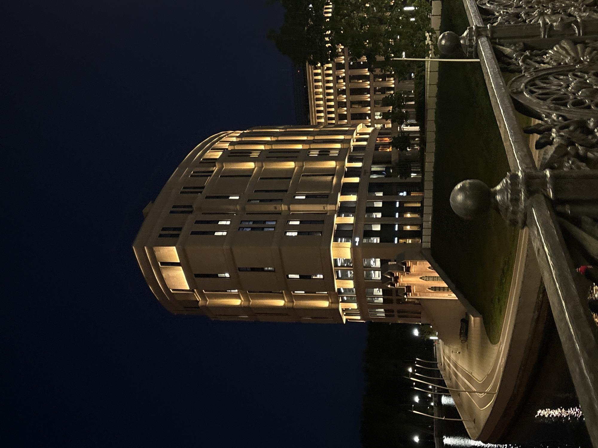 The image shows a night view of a building in Minsk, Belarus, illuminated with golden lights. The building has a curved facade with multiple windows and is likely a commercial or residential structure. In the foreground, a bridge railing with decorative metalwork is visible. To the right of the building, there is a green area, possibly a park or lawn. The lighting suggests an urban setting with streetlights and other buildings in the background, adding to the cityscape.