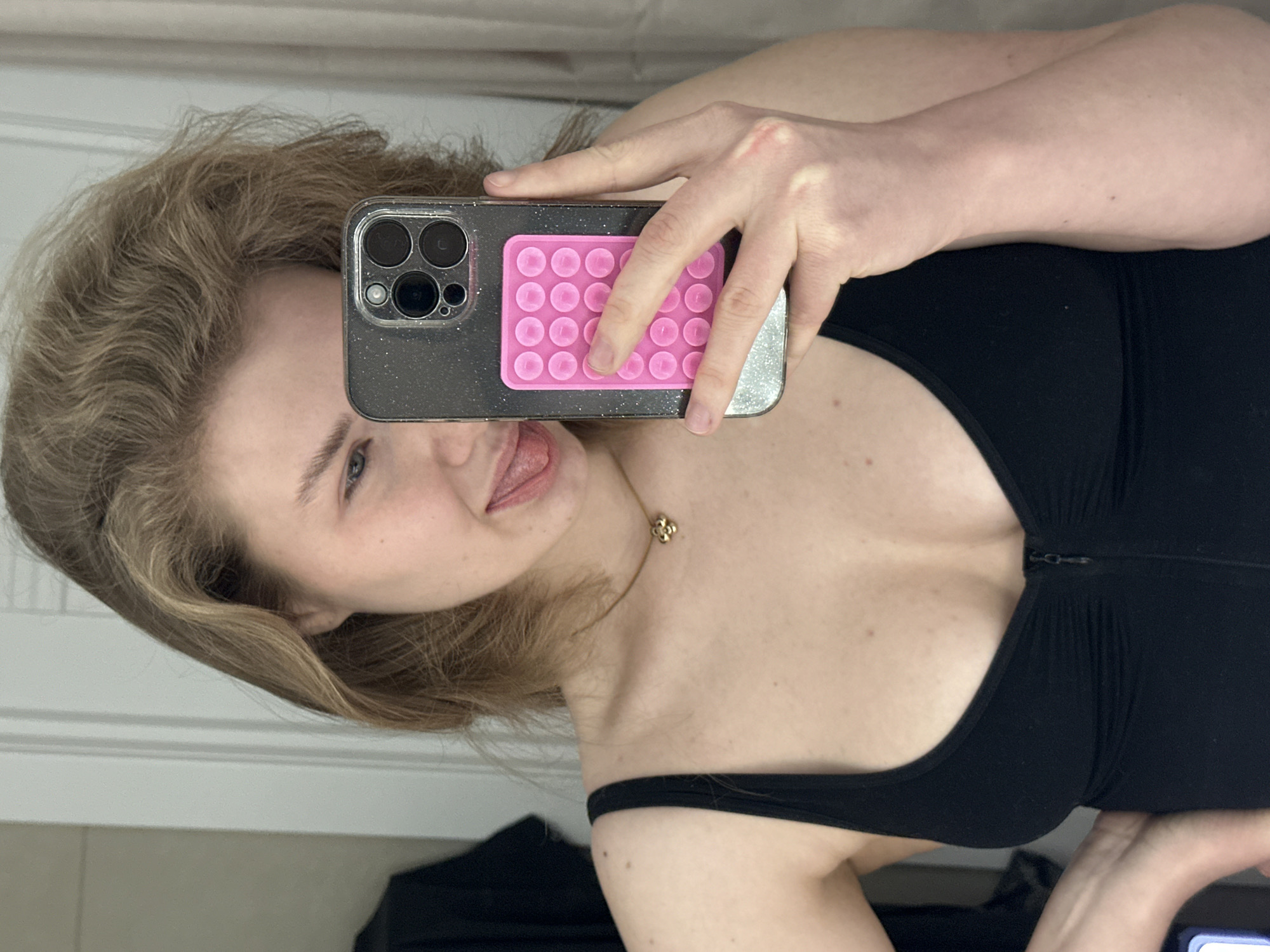 A light-skinned, young woman is taking a selfie in front of a mirror. She is holding a dark grey smartphone with a pink pop-it attached to the back. She is wearing a black top and a gold necklace. The background suggests an indoor setting with white walls and some visible clothing. The location in Kazan, Russia, is unknown.