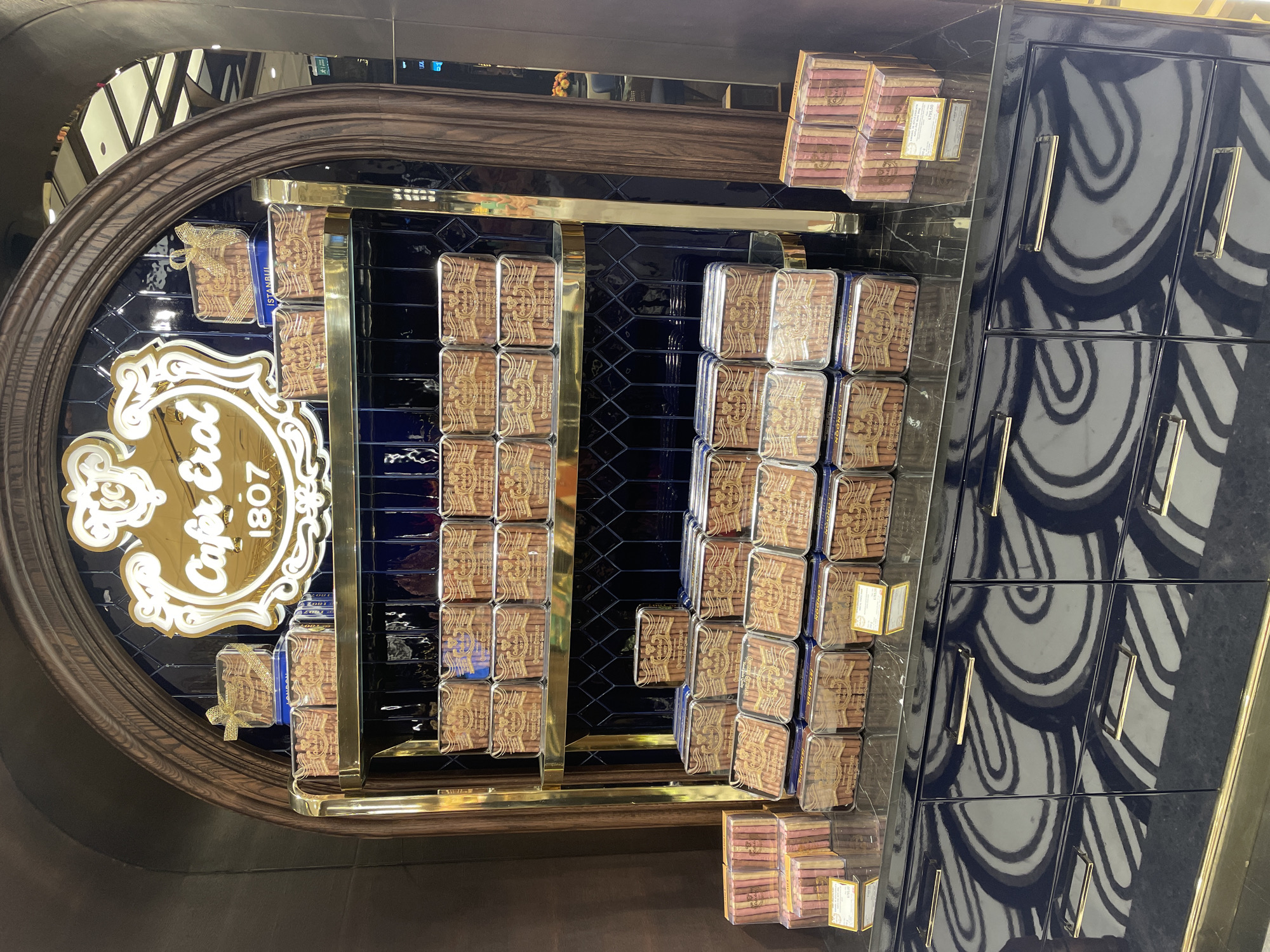 The image shows a display of luxury confectionery in what appears to be a retail establishment. The products are neatly arranged on gold shelves against a dark blue tiled wall, framed by a dark wooden arch. A large, ornate sign above the display reads "Cafer Erol 1807," suggesting a historical brand. The confectionery is packaged in rectangular tins, many of which feature detailed gold lettering and imagery. Some tins are stacked, while others are presented individually. On the lower shelf, there are stacks of what appear to be biscuit-like items in pink and brown packaging. The overall presentation is elegant and suggests high-quality goods. The reflective surface of the counter below the shelves shows a swirling pattern, and the cabinetry underneath is a deep blue with gold handles. There are no people visible in the image. The reference to Kadıköy, Turkey, strongly suggests this is a display from Cafer Erol, a well-known Turkish confectionery shop located in Kadıköy, Istanbul.