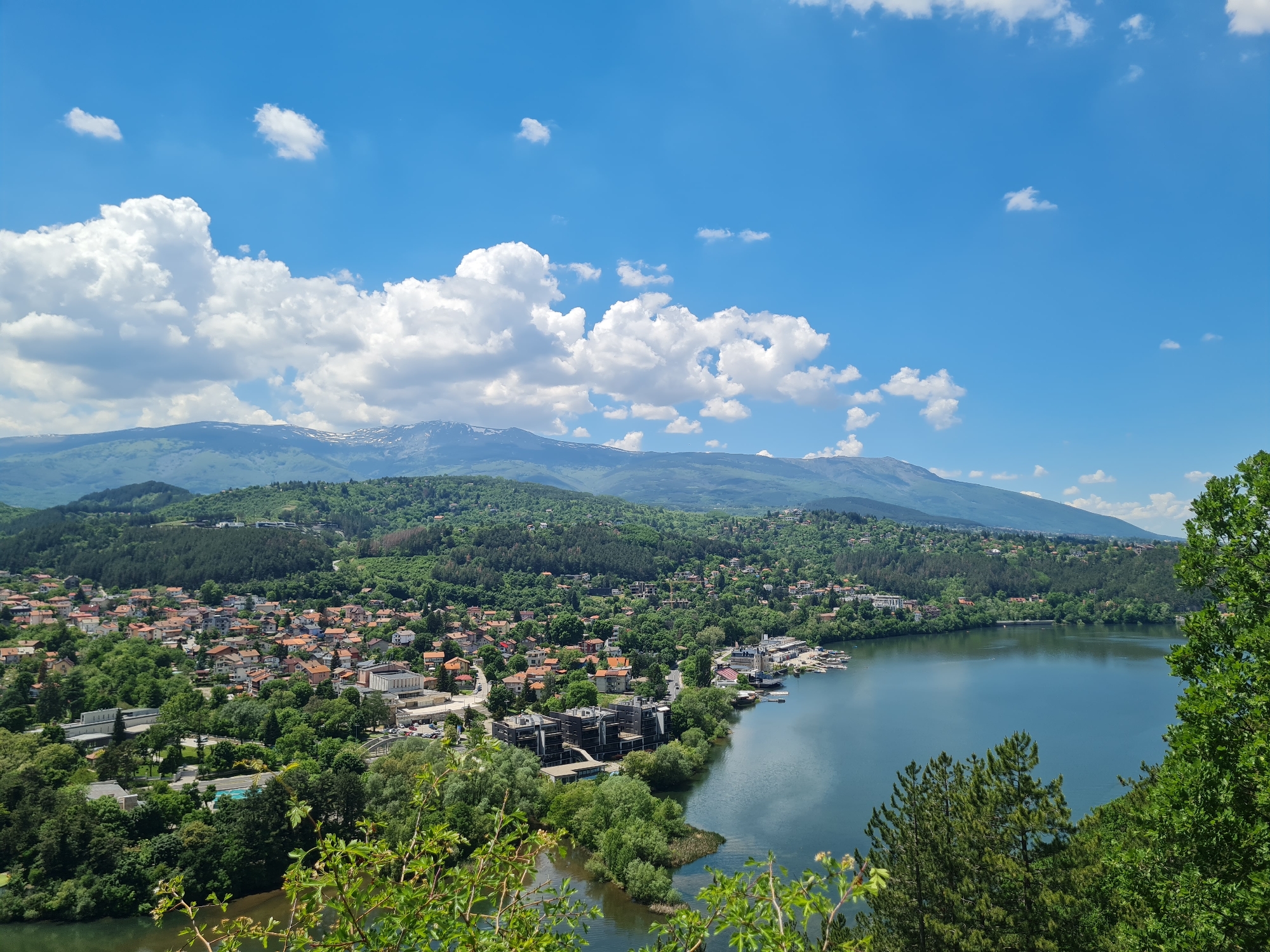 The image presents a high-angle view of Pancharevo Lake and a section of Pancharevo, Bulgaria. The scene is dominated by a large lake occupying the right half of the frame. A section of the town, characterized by houses with orange roofs, is nestled amongst dense green trees to the left of the lake. The surrounding hills are also heavily forested. In the background, mountains are visible under a bright blue sky dotted with fluffy white clouds.